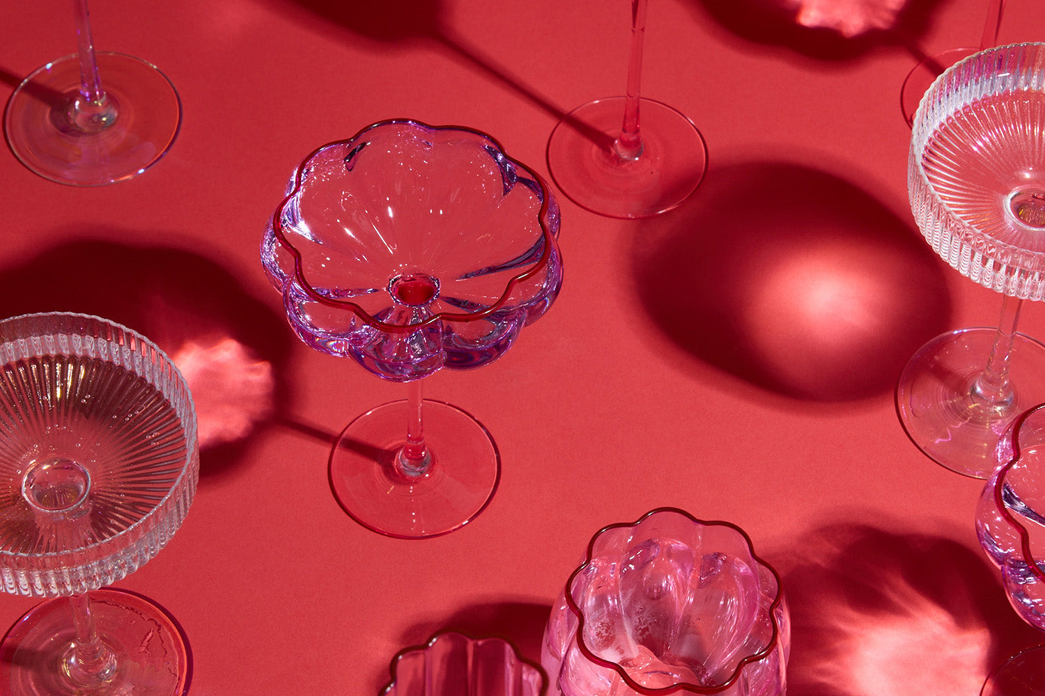 A group of empty, decorative glassware with intricate patterns casts colorful shadows on a bright red surface. The glass stems and bowls create playful reflections and shapes.