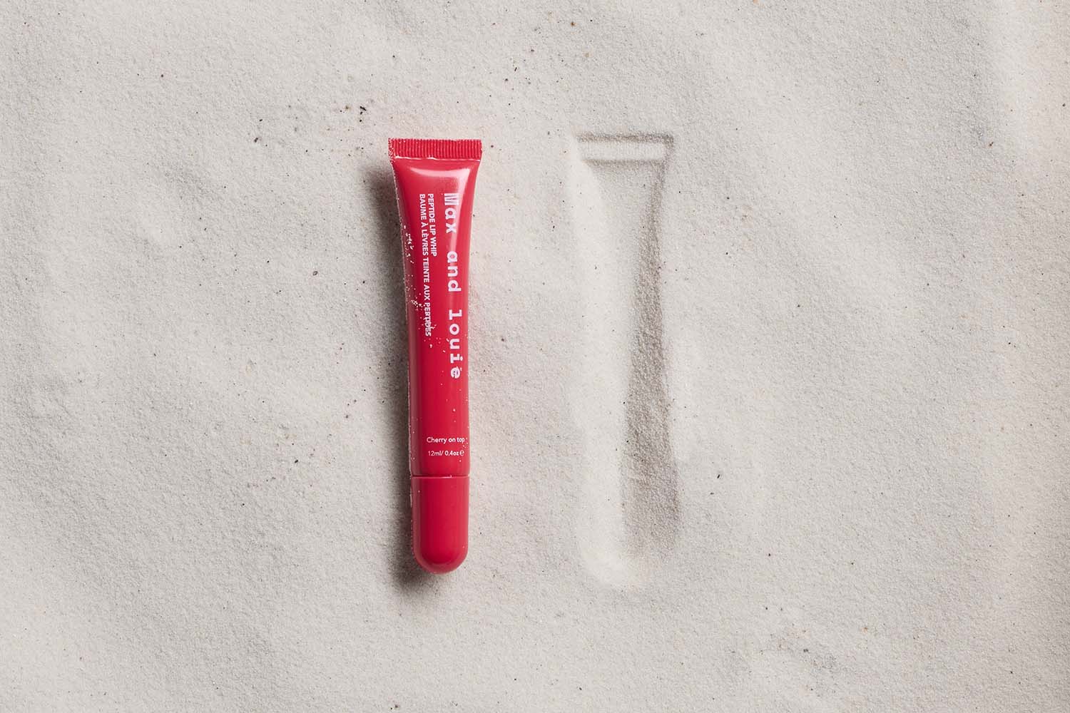 A red tube of lip balm labeled Wax and Lous lies on white sand next to its imprint, showing where it was placed earlier.