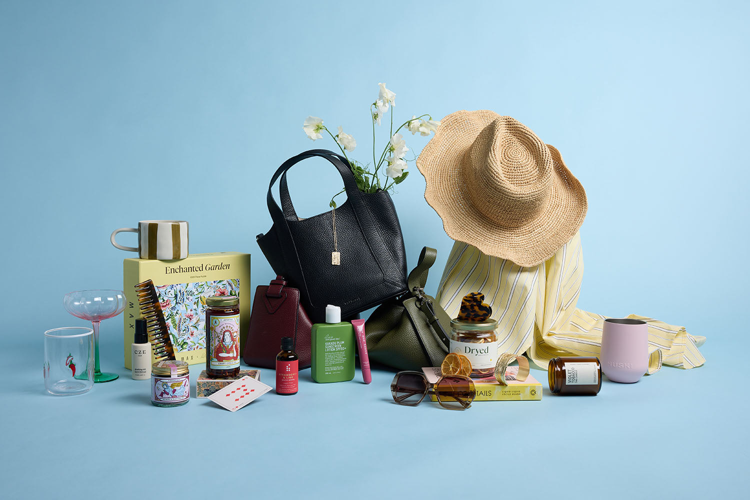 A collection of gifts on a blue background, including handbags, a straw hat, sunglasses, mugs, beauty products, a striped shirt, flowers, a candle, playing cards, and a book.