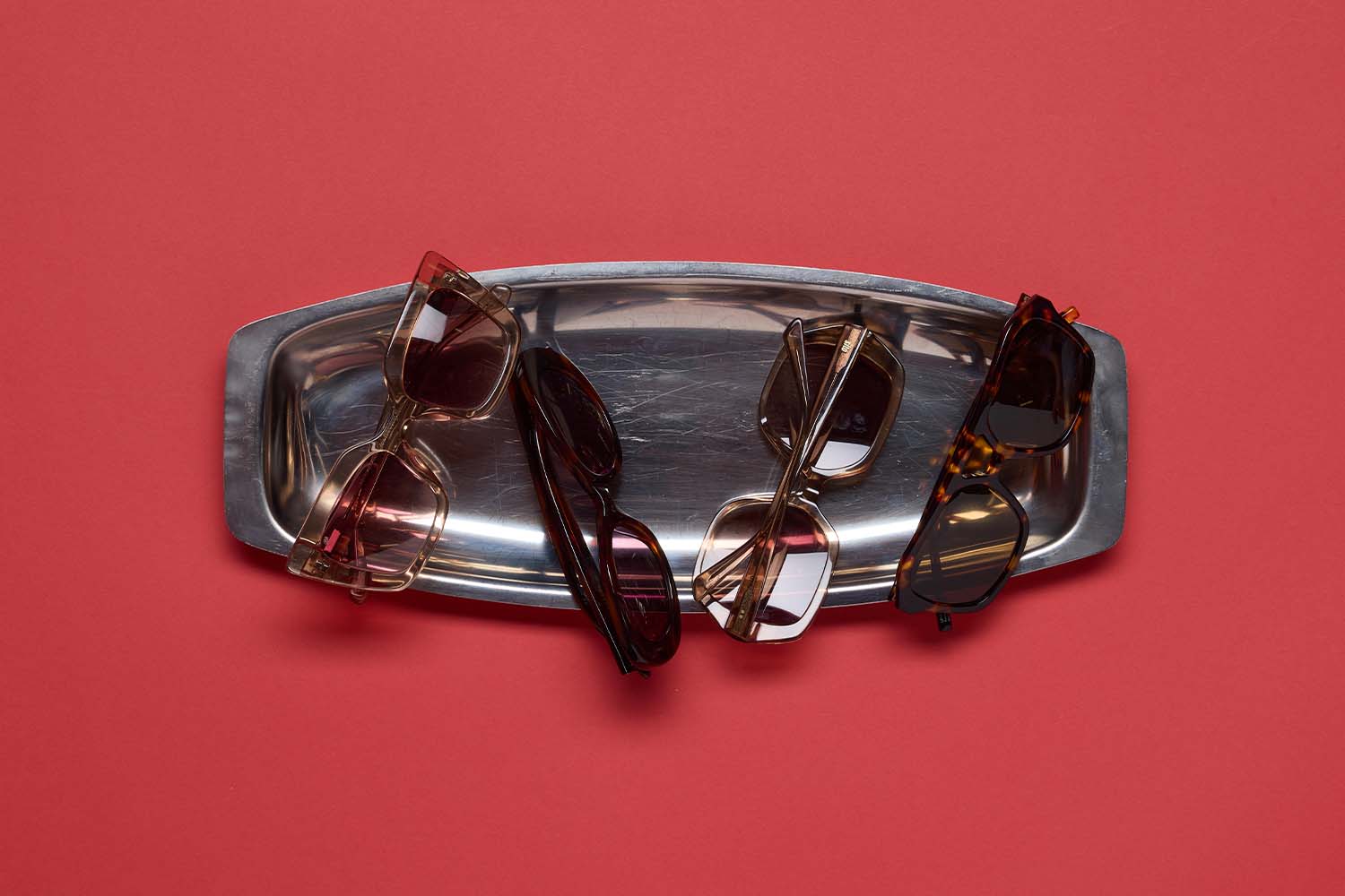A silver tray holds five pairs of stylish sunglasses in various colors and shapes, displayed on a red background.