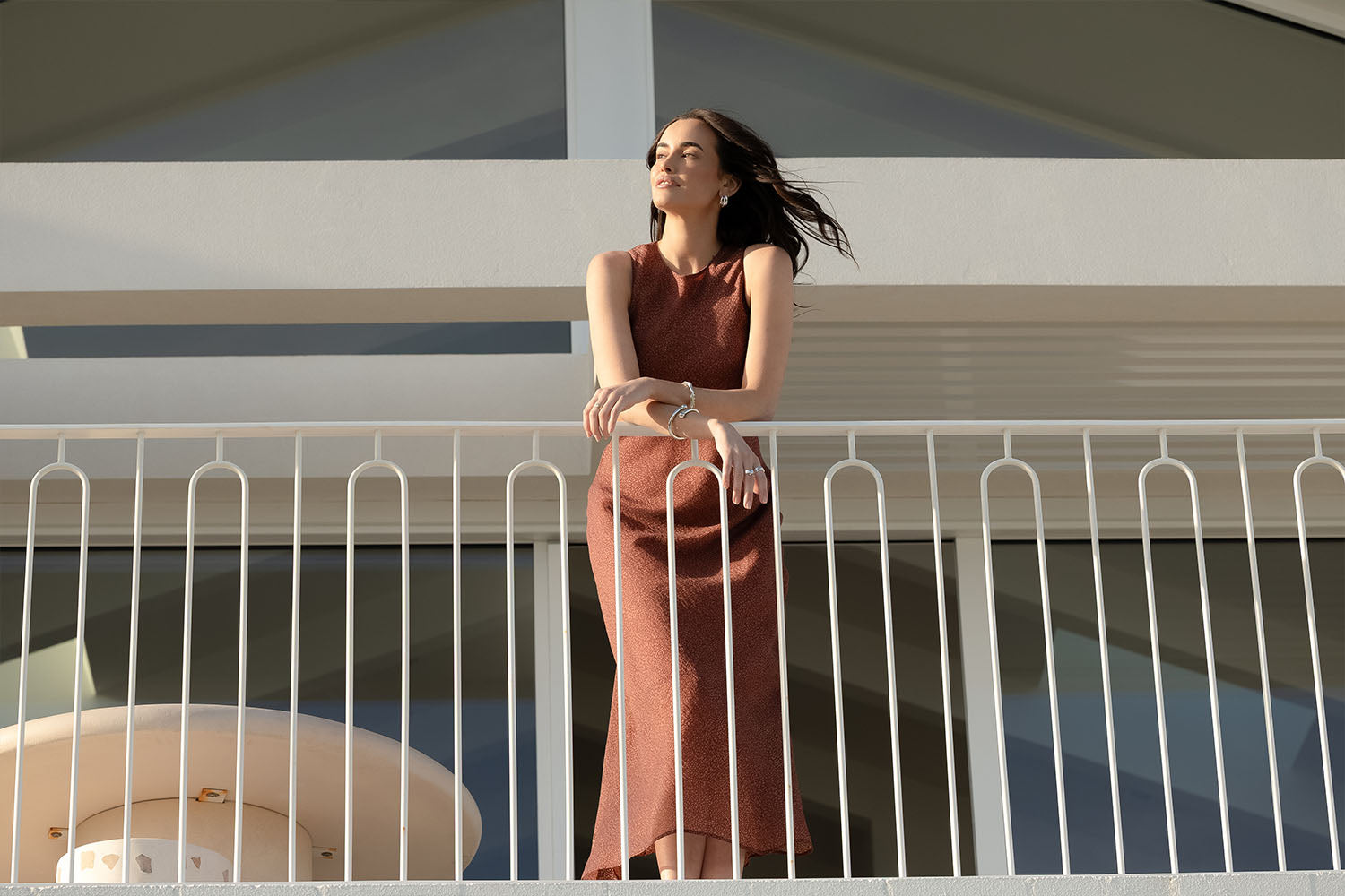 A woman in a sleeveless brown dress leans on a white balcony railing, eyes closed, with sunlight illuminating her face. Modern architectural elements are visible in the background.