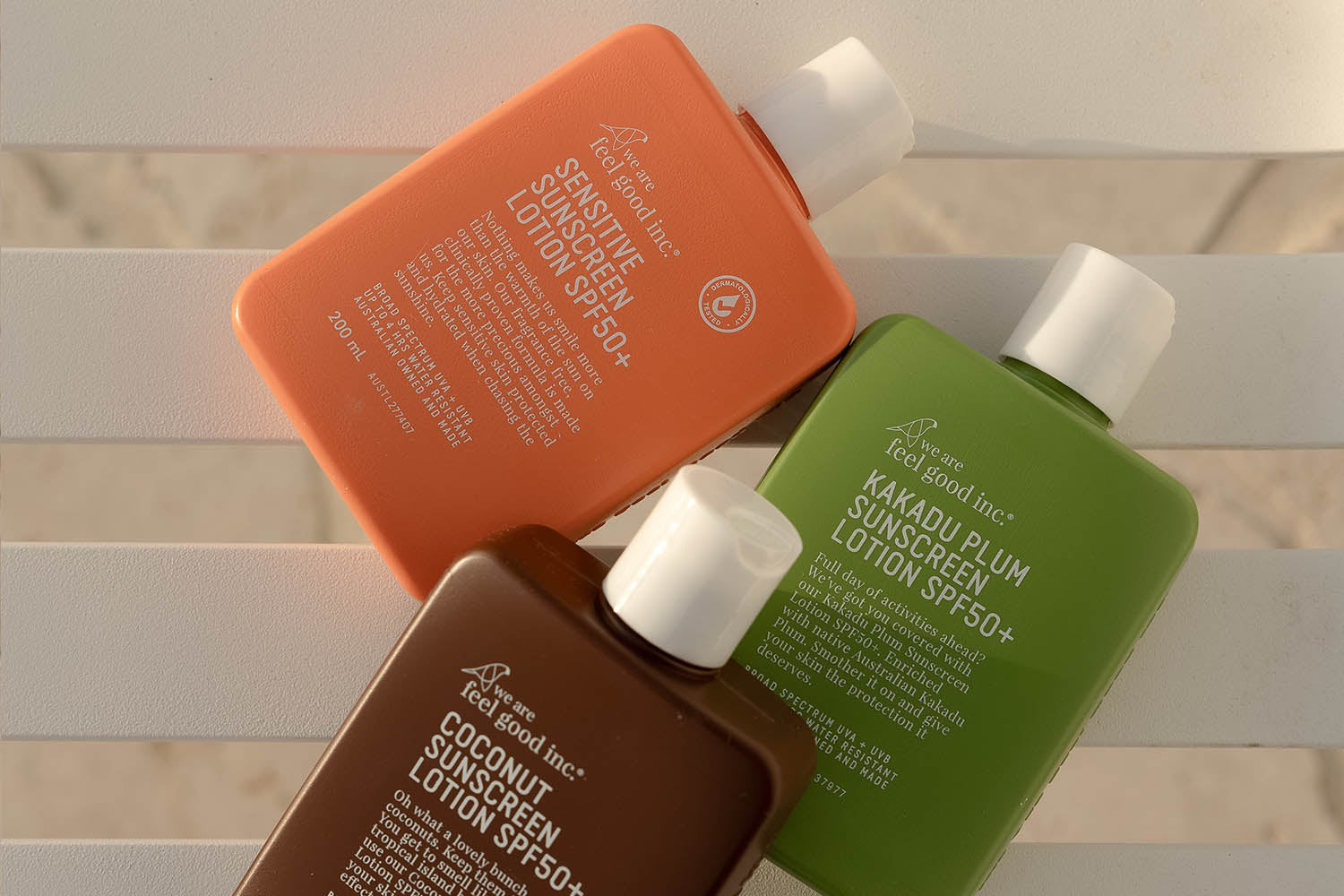 Three rectangular bottles of Feel Good Inc. sunscreen lotion in orange, green, and brown are placed on a white slatted surface, lit by natural sunlight.