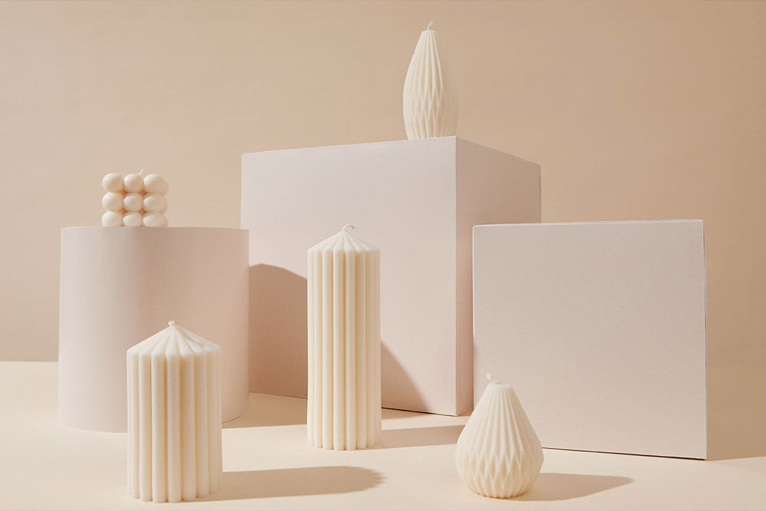 Five cream-colored, uniquely shaped candles are arranged on and around geometric white blocks against a beige background, creating a minimalist and modern aesthetic.