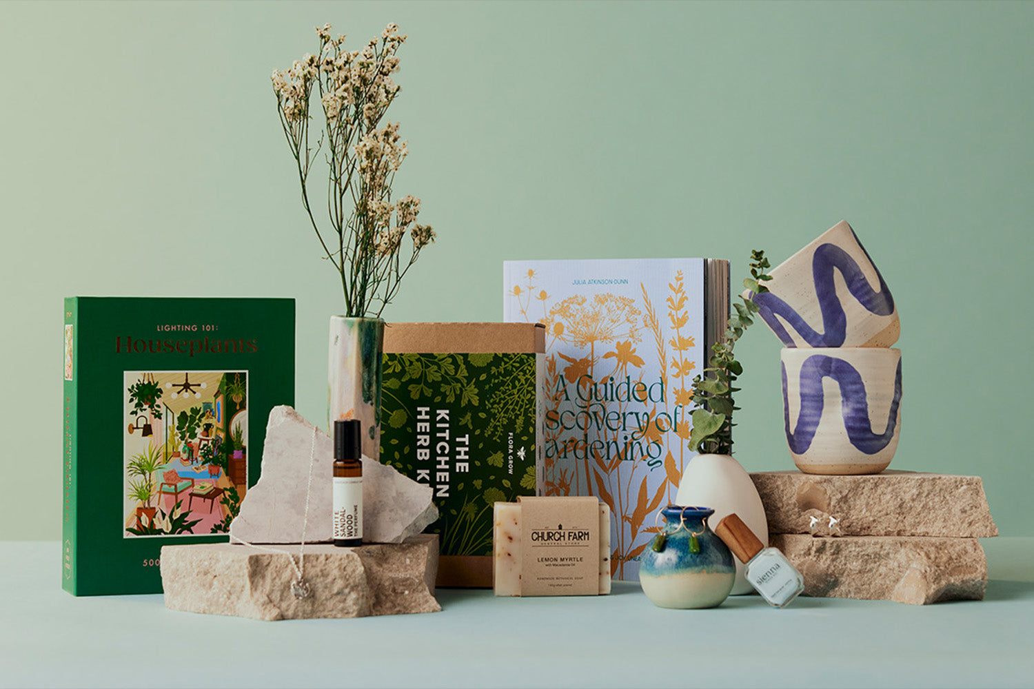 A collection of books, ceramics, a small vase, skincare products, and dried flowers artfully arranged on stone slabs against a pale green background.