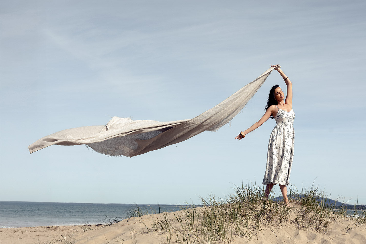 A woman in a white dress stands on a sandy beach, holding a long, flowing fabric above her head as it billows in the wind. The sky is clear and the ocean is visible in the background.