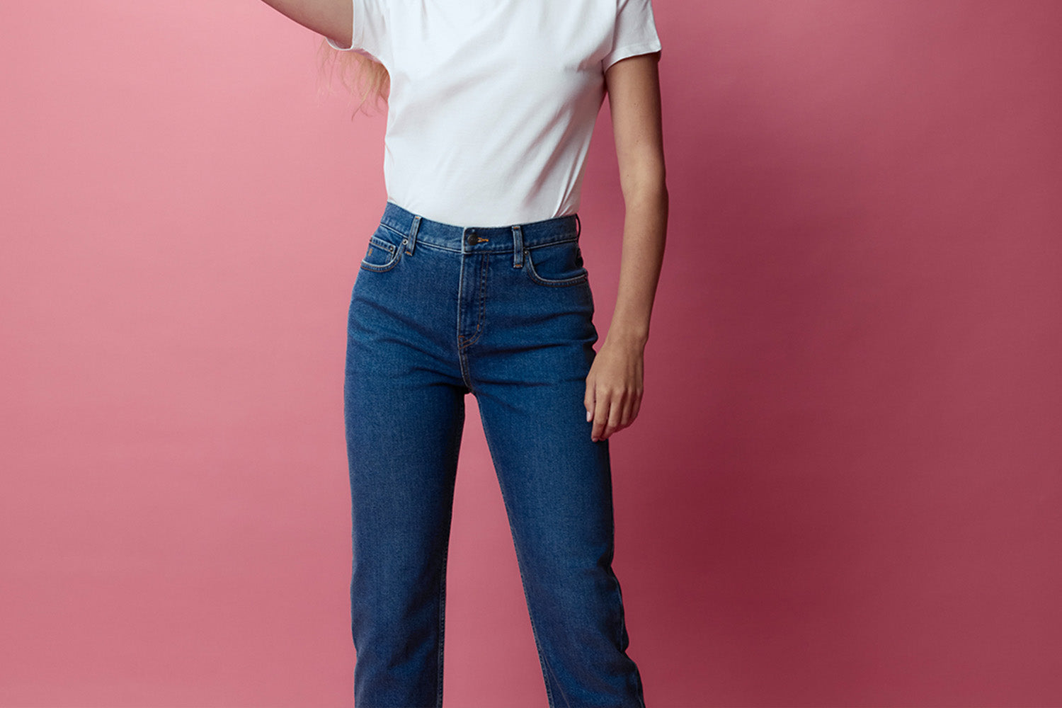 A person wearing a white t-shirt and blue jeans stands against a pink background. Their face is partially out of frame, and one arm is raised, with only the lower part of the body visible.