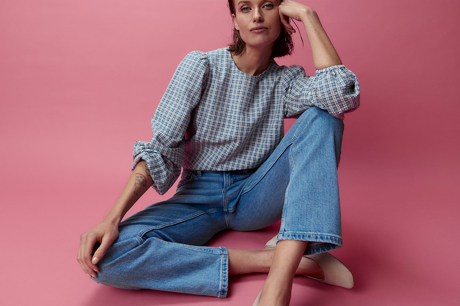 A woman in a plaid blouse and blue jeans sits on a pink floor with one leg bent and her elbow resting on her knee, posing against a pink background.