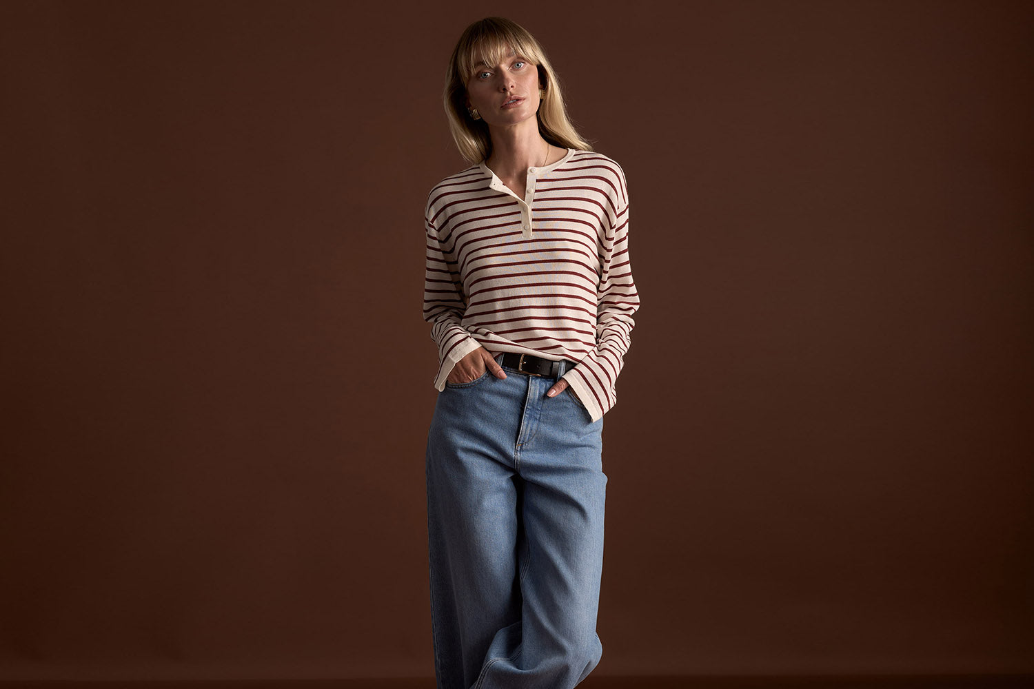 A woman stands against a brown background, wearing a long-sleeve, cream-colored shirt with red and brown stripes and blue high-waisted jeans. She has blonde hair with bangs and one hand in her pocket.