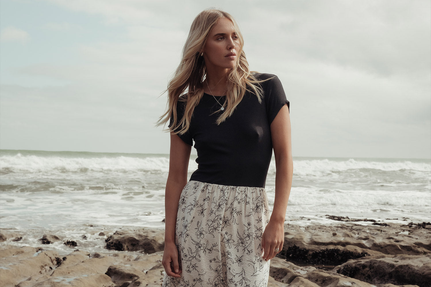 A woman with long blonde hair stands on a rocky beach wearing a black short-sleeve top and a high-waisted, white skirt with a floral pattern. Waves and a cloudy sky are in the background.