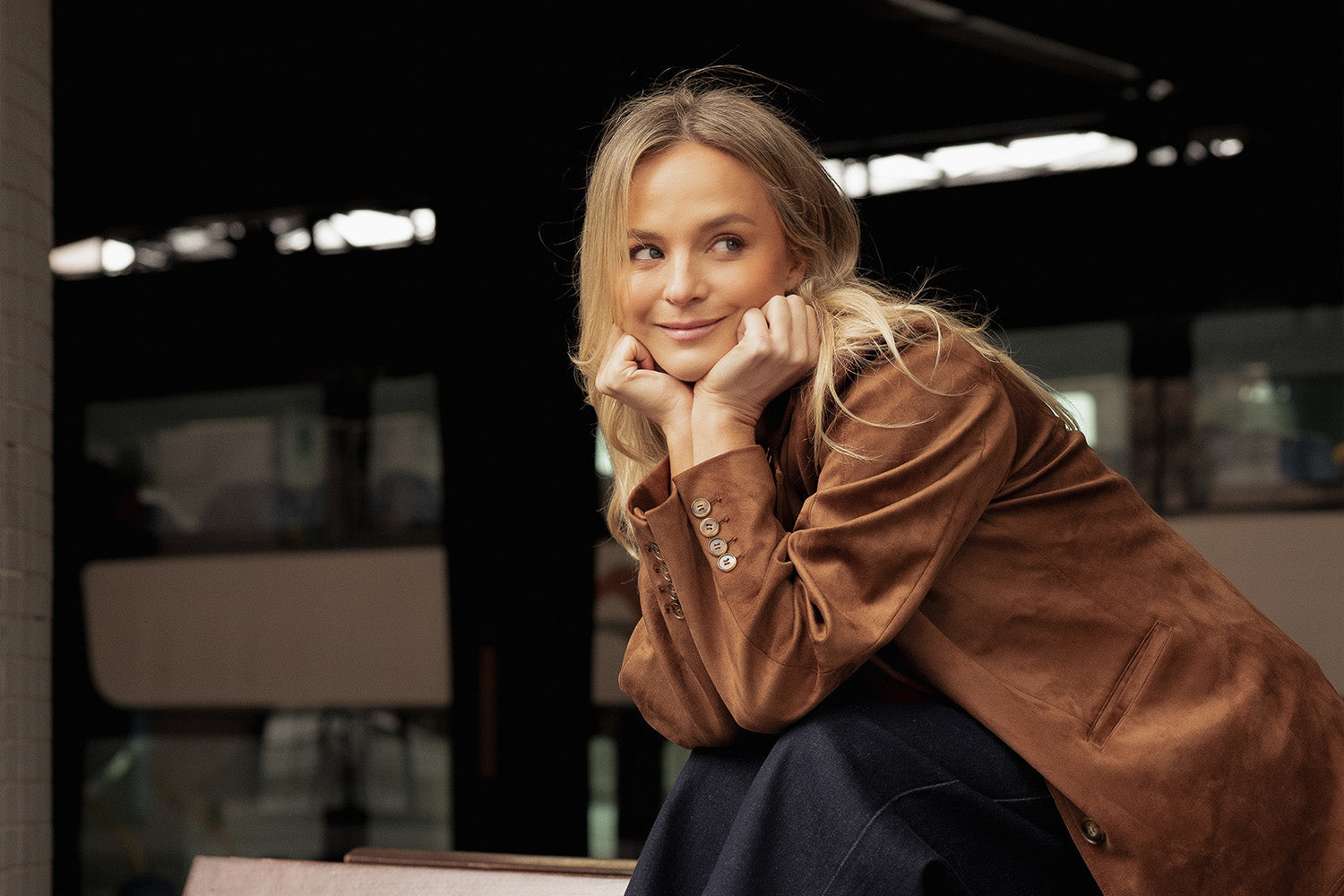 A woman with long blonde hair and a brown suede jacket sits with her chin resting on her hands, smiling and looking to the side. The background is dark and out of focus.