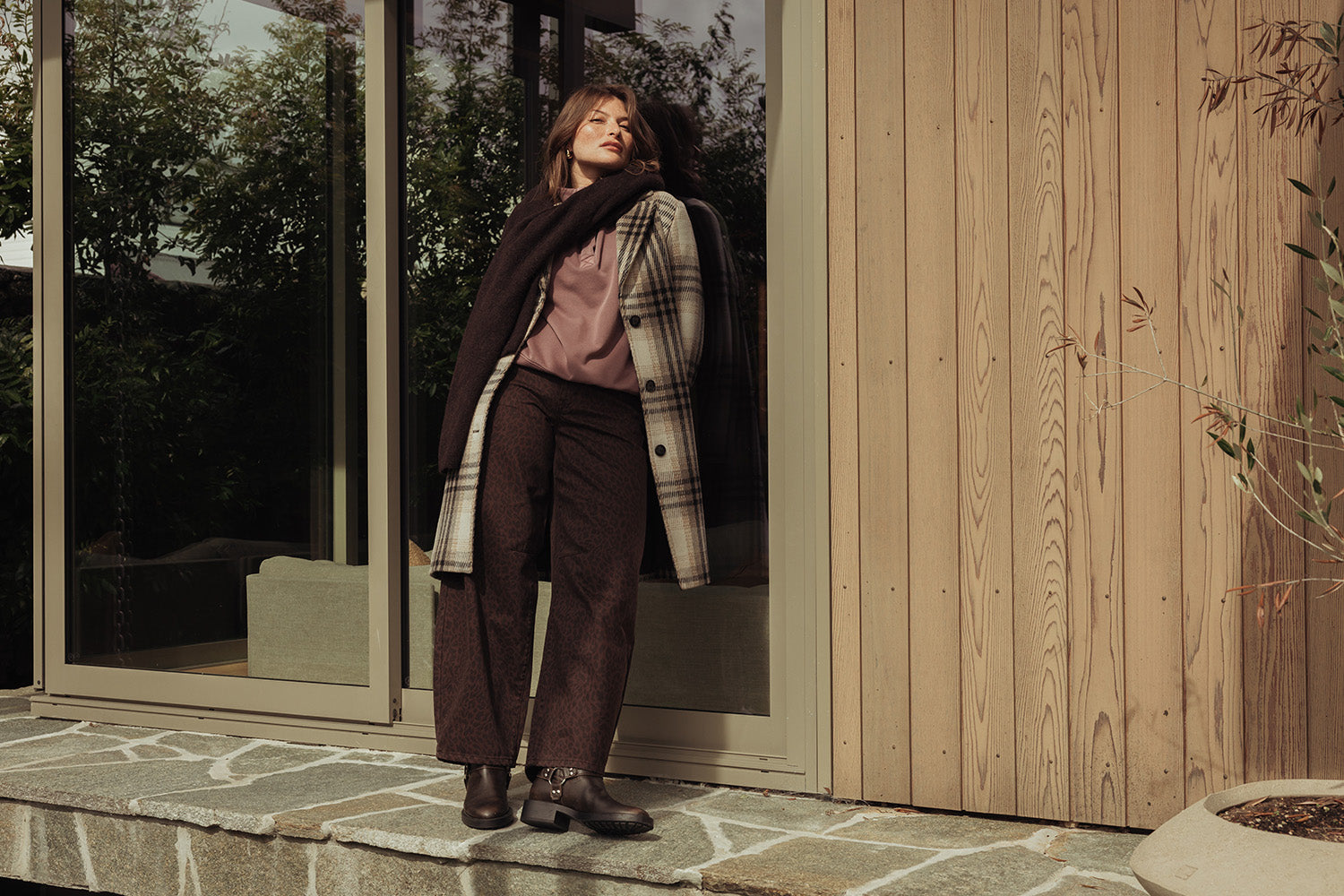A woman in a plaid coat, mauve blouse, and dark pants stands on a stone porch, leaning against a wooden wall near large glass doors, looking thoughtfully into the distance.