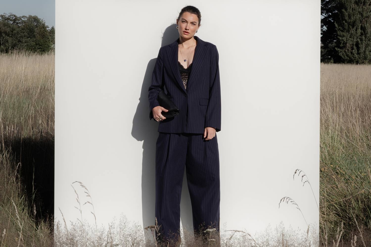 A woman in a plaid coat, mauve blouse, and dark pants stands on a stone porch, leaning against a wooden wall near large glass doors, looking thoughtfully into the distance.