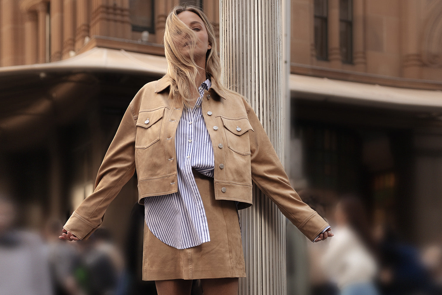A woman stands outdoors with wind blowing her hair over her face, wearing a tan suede jacket and skirt over a blue-and-white striped shirt. The background shows an urban setting with blurred people.