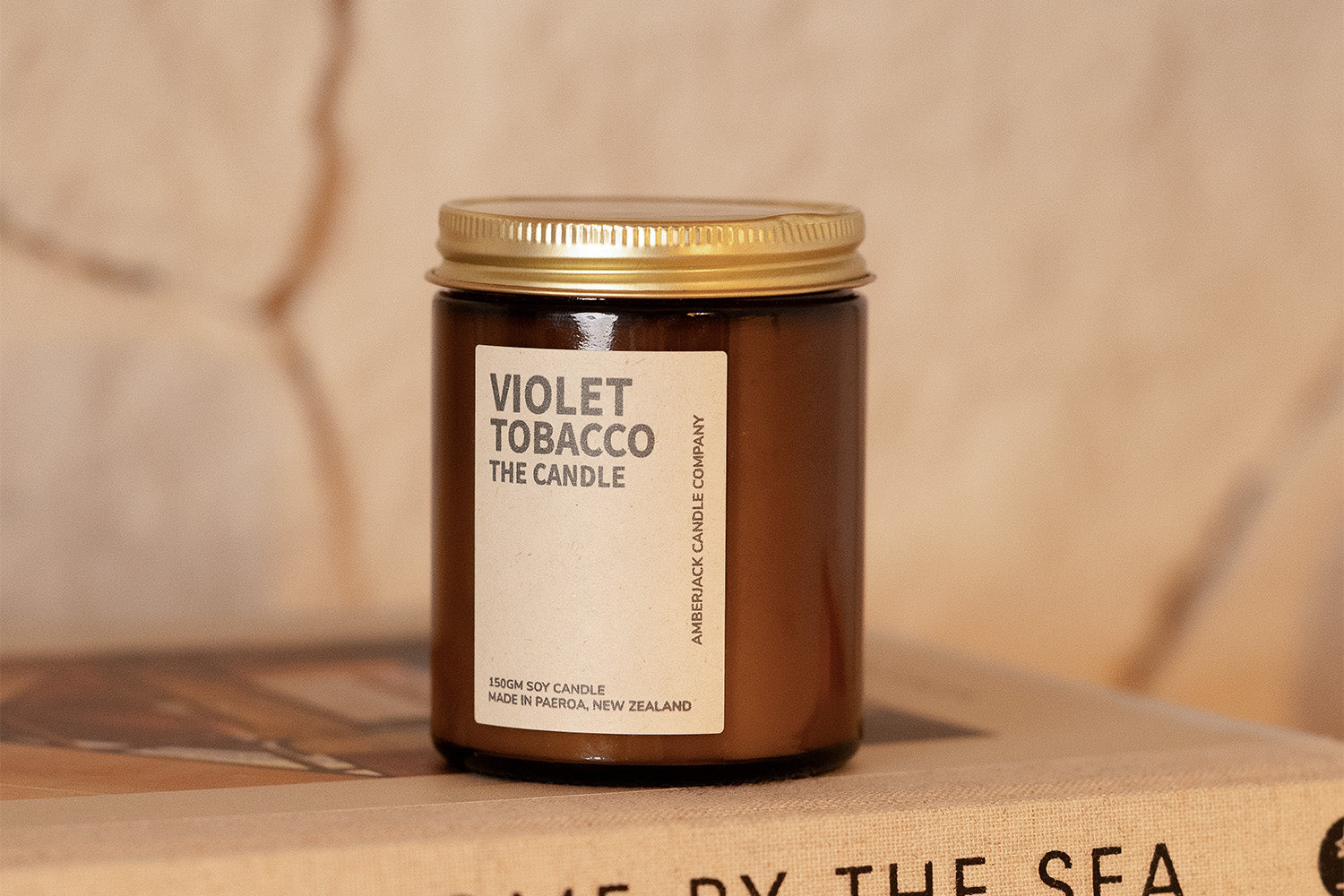 A brown glass jar candle labeled Violet Tobacco The Candle with a gold lid sits on a closed book. The label notes its a 250ml soy candle made in Pareora, New Zealand.