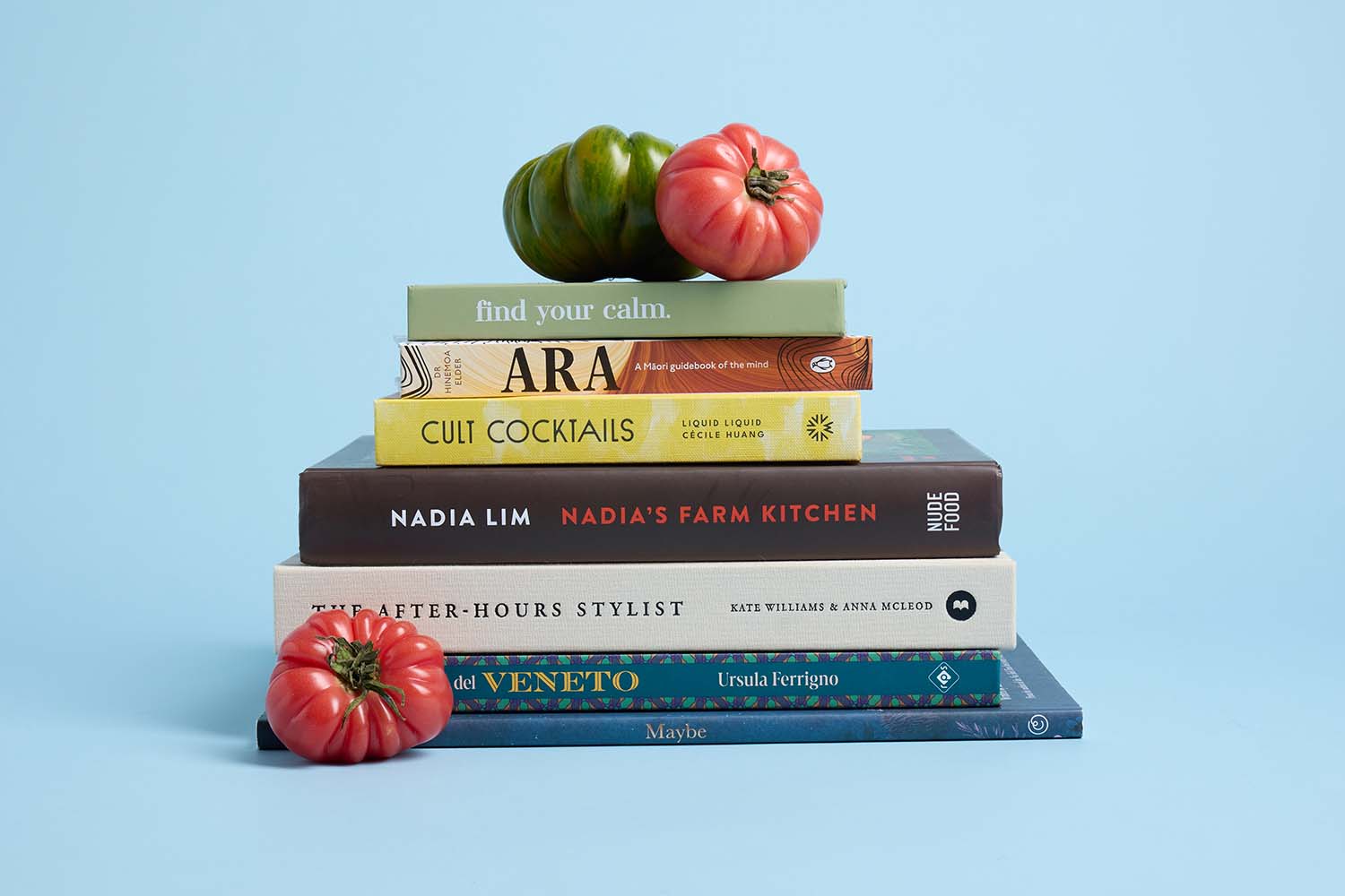A stack of seven books and three heirloom tomatoes are arranged against a light blue background. Some books include titles like Find Your Calm and Nadias Farm Kitchen.