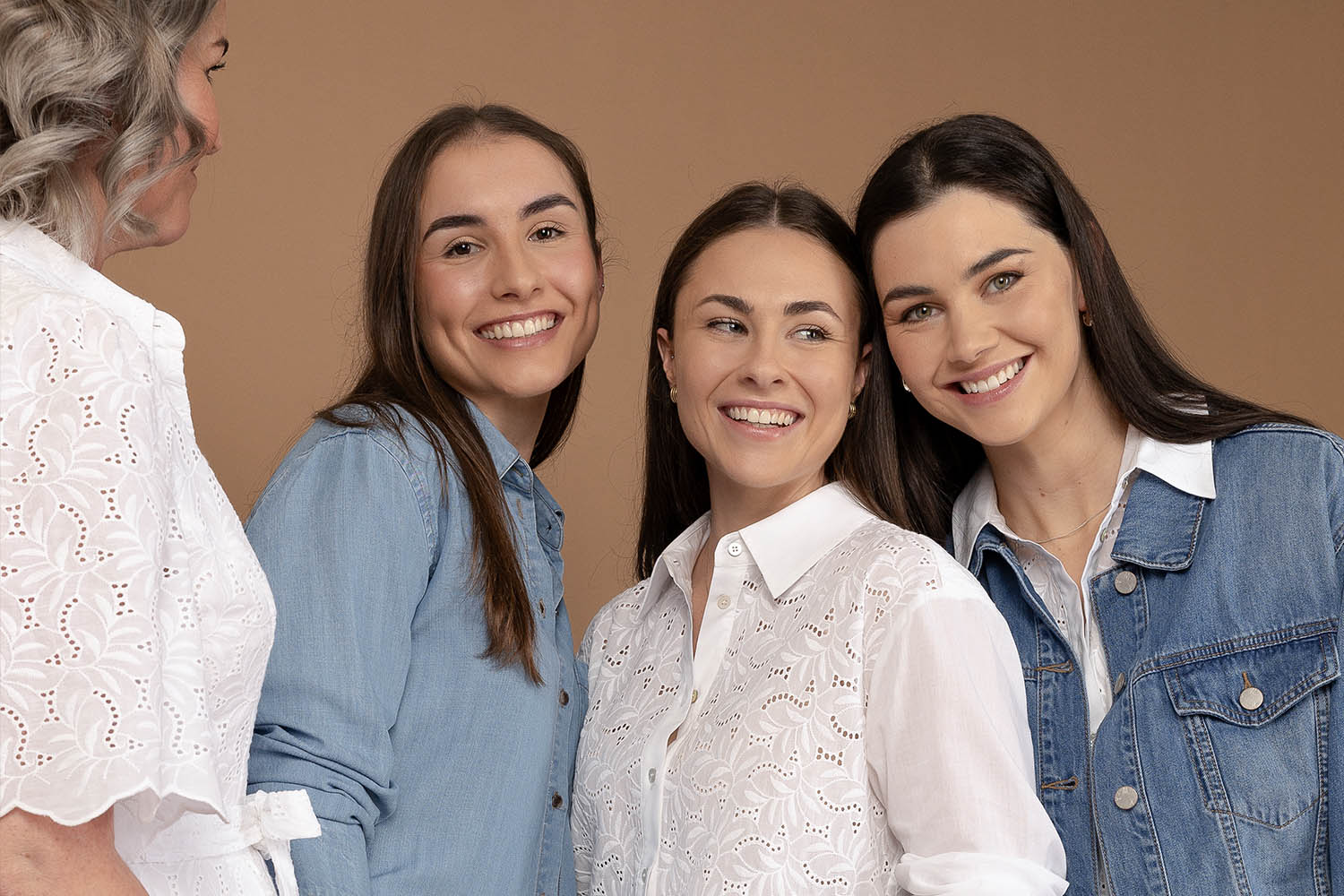 Four women stand together smiling. Three face the camera, wearing white or denim tops, while one, with gray hair, is turned to the side. The background is a plain warm brown.