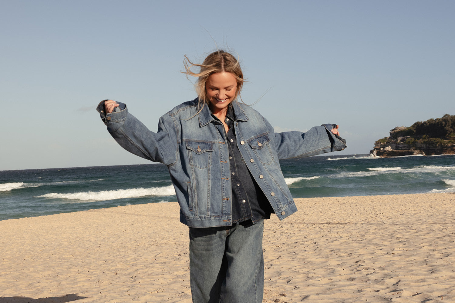 A person wearing a denim jacket and jeans smiles and spreads their arms wide while standing on a sandy beach with waves and blue sky in the background.