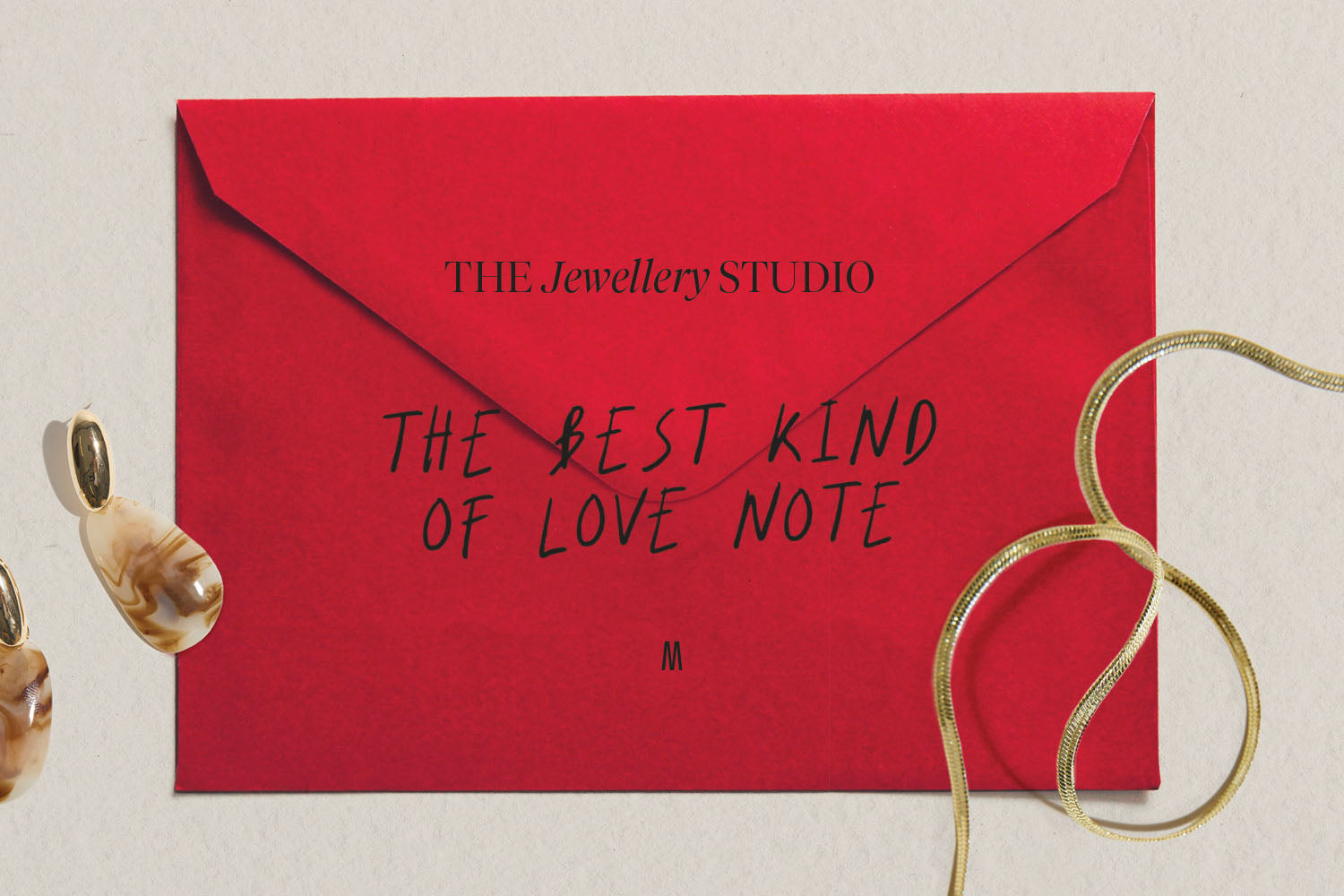 A red envelope with THE Jewellery STUDIO and THE BEST KIND OF LOVE NOTE written on it, next to a gold necklace and dangling earrings on a light surface.