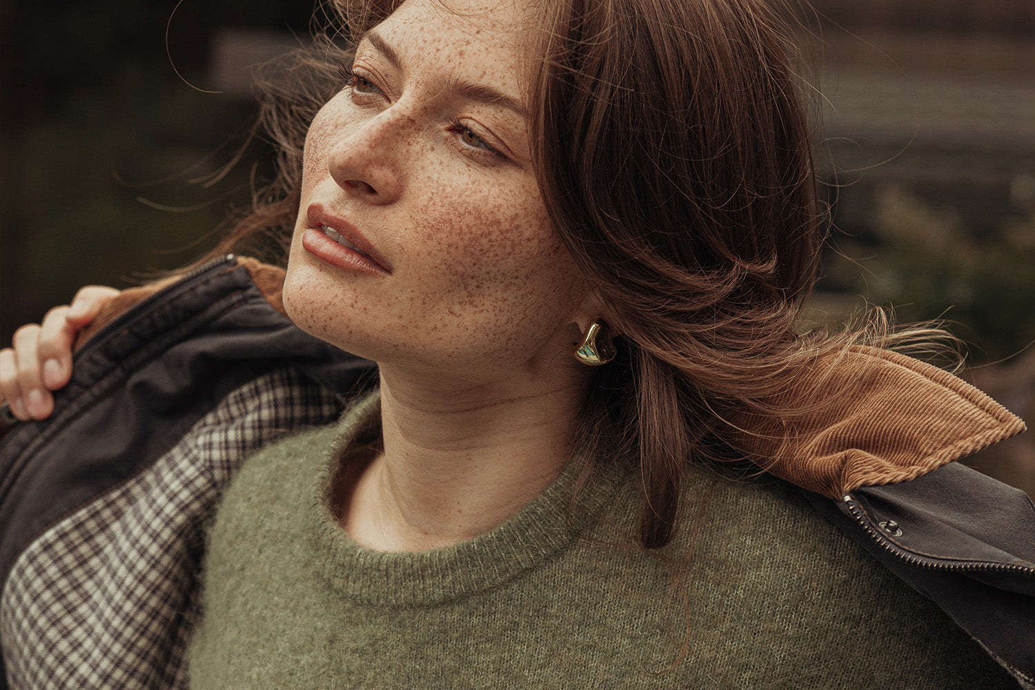 A woman with freckles and brown hair gazes into the distance, wearing a green sweater and an open jacket with a brown corduroy collar. She has one hand on her jacket and wears gold earrings.