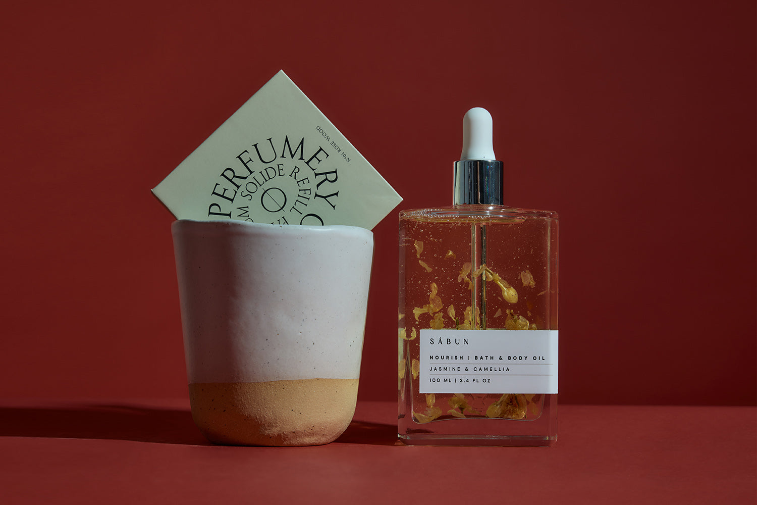 A square glass bottle of jasmine and camellia body oil with a dropper sits beside an off-white ceramic cup holding a box labeled Perfumery, against a reddish-brown background.