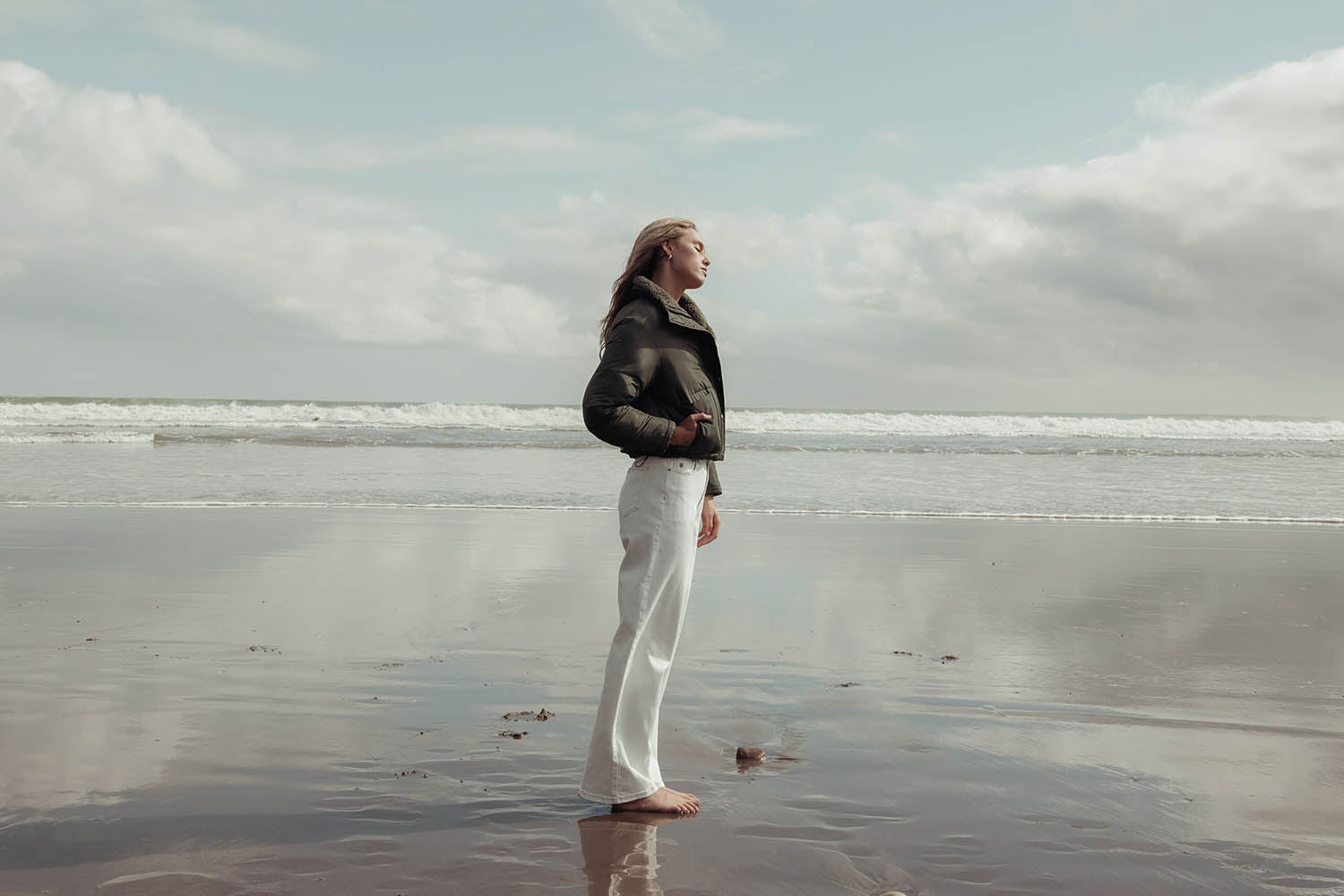 A woman stands barefoot on a wet sandy beach, wearing white pants and a dark jacket, gazing out towards the sea under a cloudy sky.