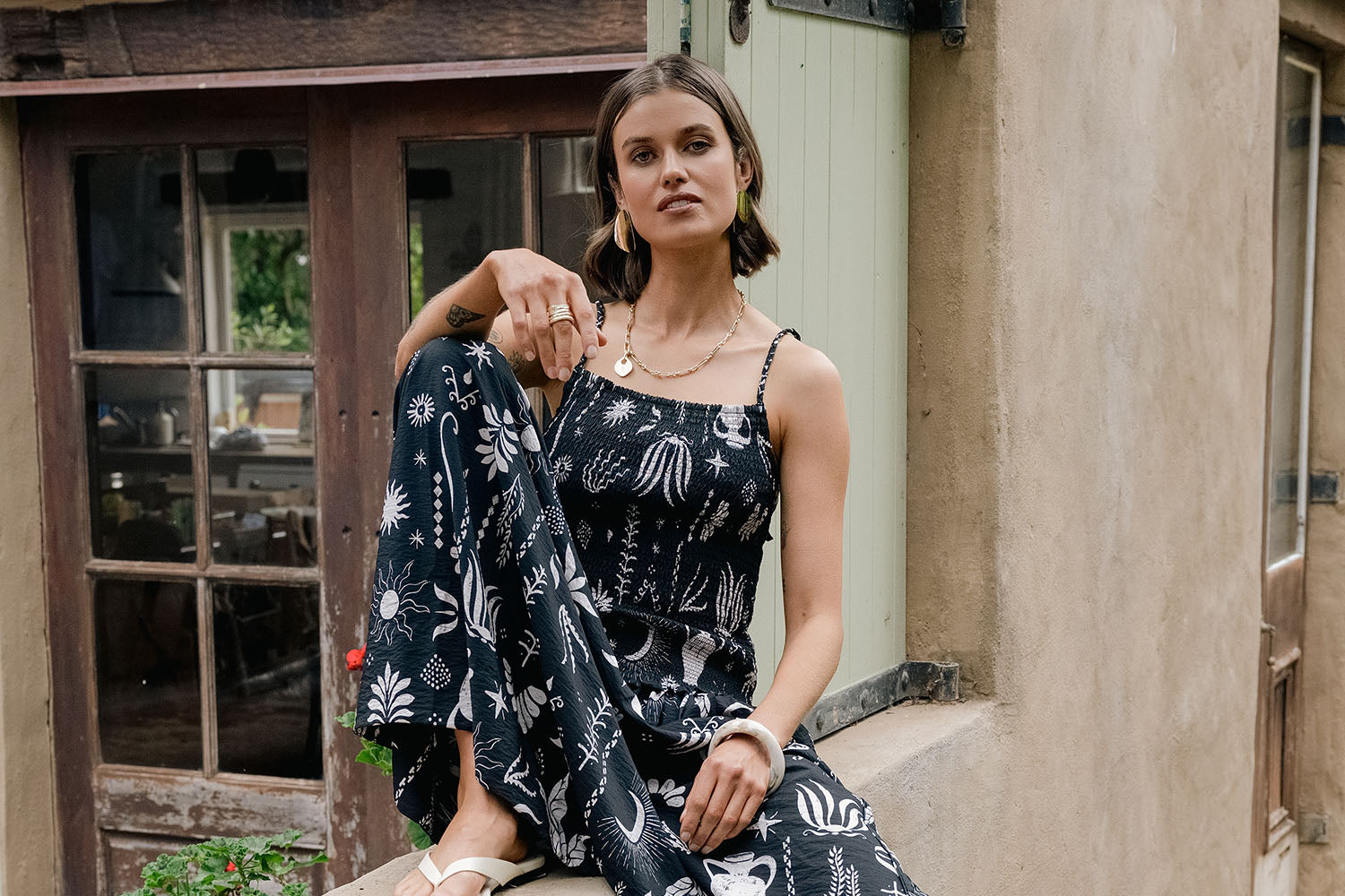 A woman in a black and white floral jumpsuit sits on a ledge outside a rustic building, looking confidently at the camera. She has short hair and wears sandals and jewelry.