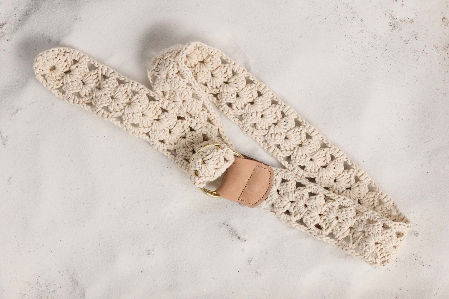 A cream-colored, crocheted belt with a brass buckle and tan leather accent is laid out on a white textured surface.