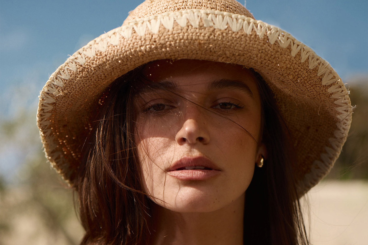 A woman with long brown hair wearing a woven straw hat looks directly at the camera; sunlight highlights her face and a few loose strands of hair blow across her face.