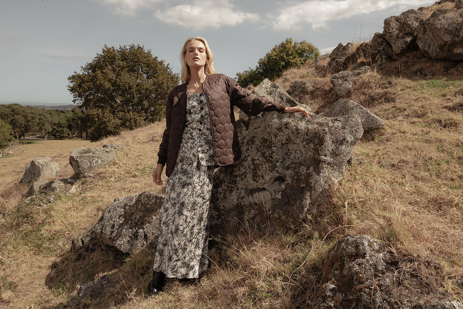 A woman in a printed maxi dress and brown quilted jacket leans on a large rock in a grassy, rocky outdoor landscape under a partly cloudy sky.