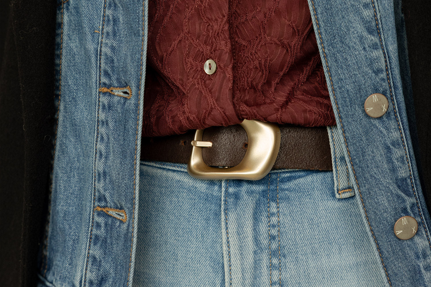 A person wearing a maroon lace shirt, blue denim jacket, blue jeans, and a dark brown belt with a large gold buckle. The image focuses on the midsection and outfit details.