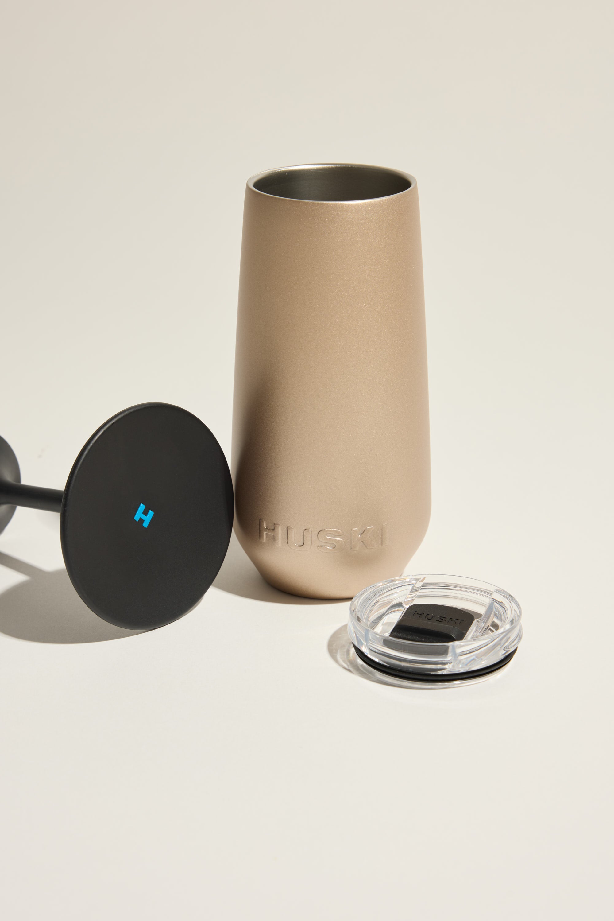 The Huski Champagne Flute by Huski, shown in matte gold with a clear lid and black round accessory, features the brand embossed on the side and is set against a light background.