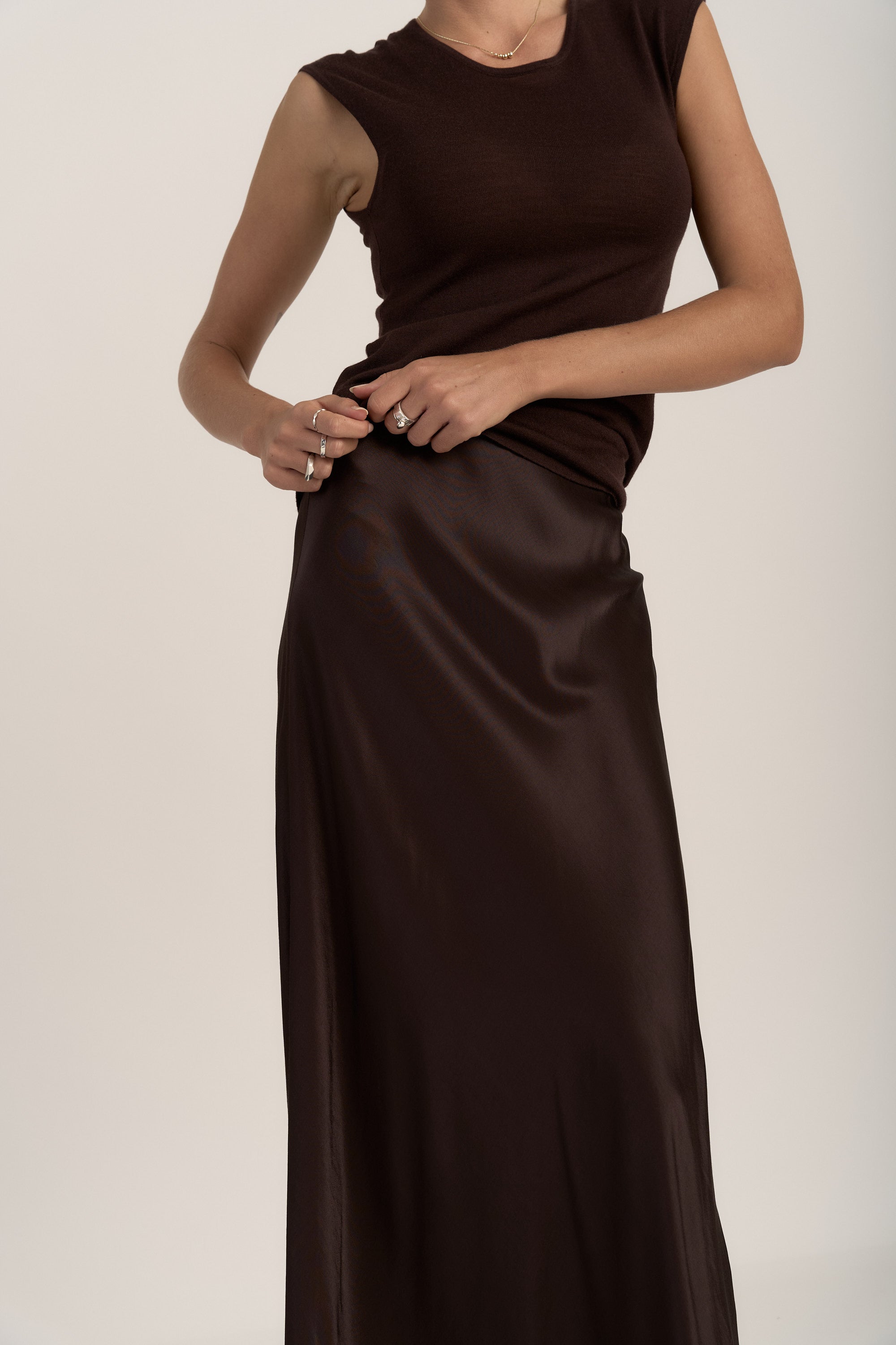 A person wearing a sleeveless dark brown top and The Slip Skirt by Max stands against a plain light background, holding the side of the shiny satin midi skirt. Their face is not visible.