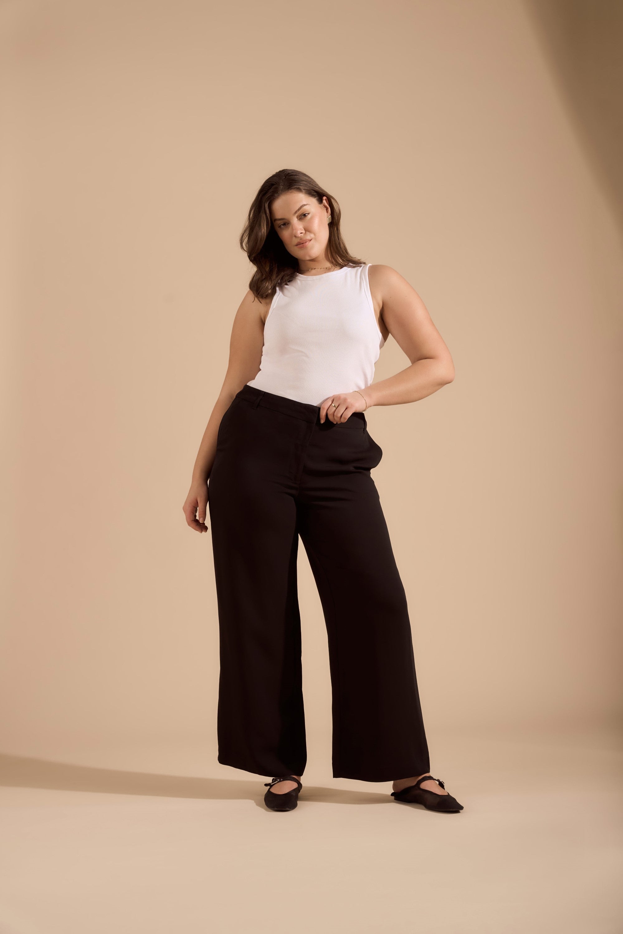 A woman with shoulder-length brown hair poses against a beige background, wearing Max’s The Wide Leg Pant in black and a white sleeveless top, standing confidently with one hand on her hip as she faces the camera.