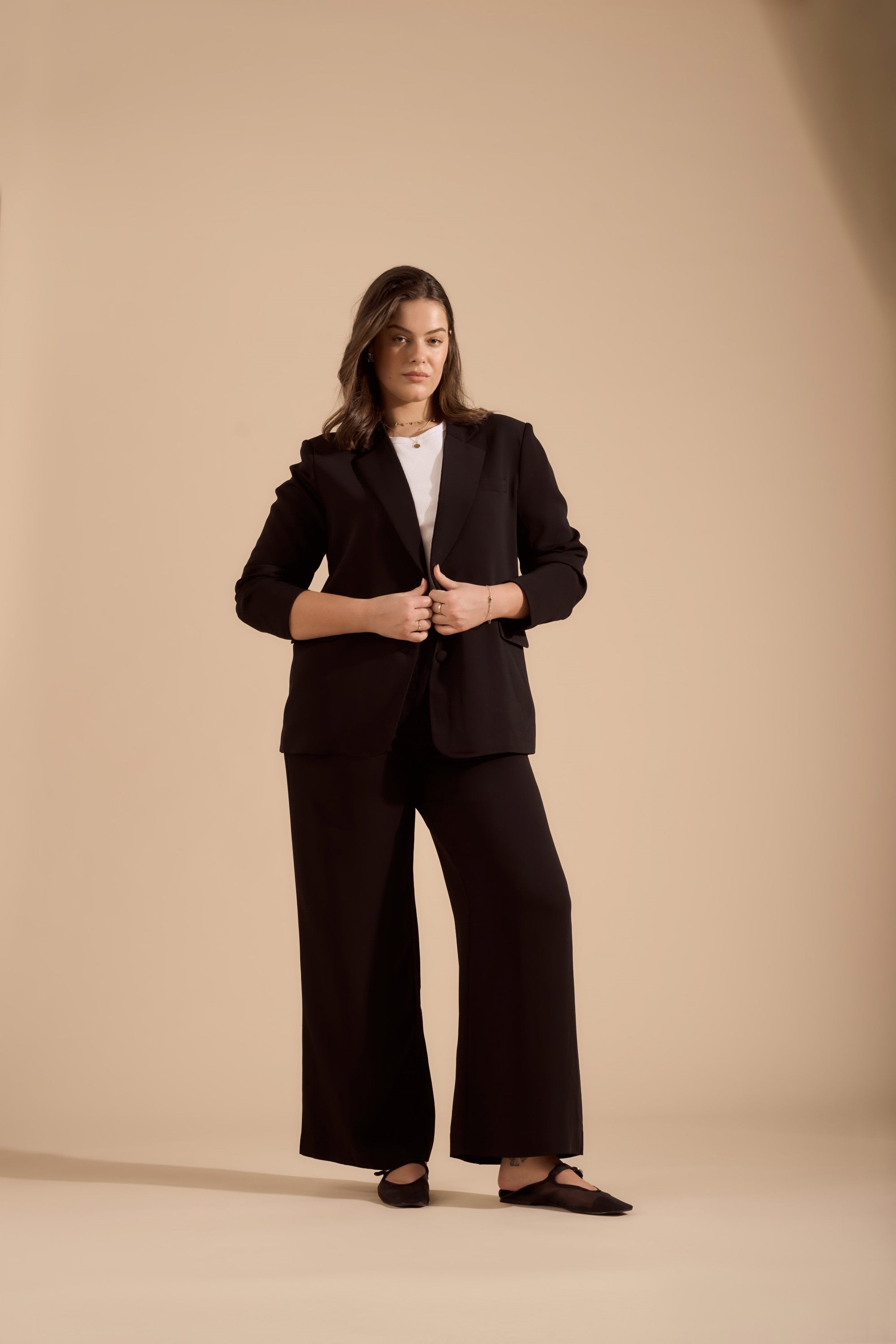 A woman stands confidently against a beige background, wearing The Wide Leg Pant by Max—a black viscose blend suit with high waisted wide leg pants, a white top, and black shoes. She has shoulder-length hair and holds her blazer’s lapels.