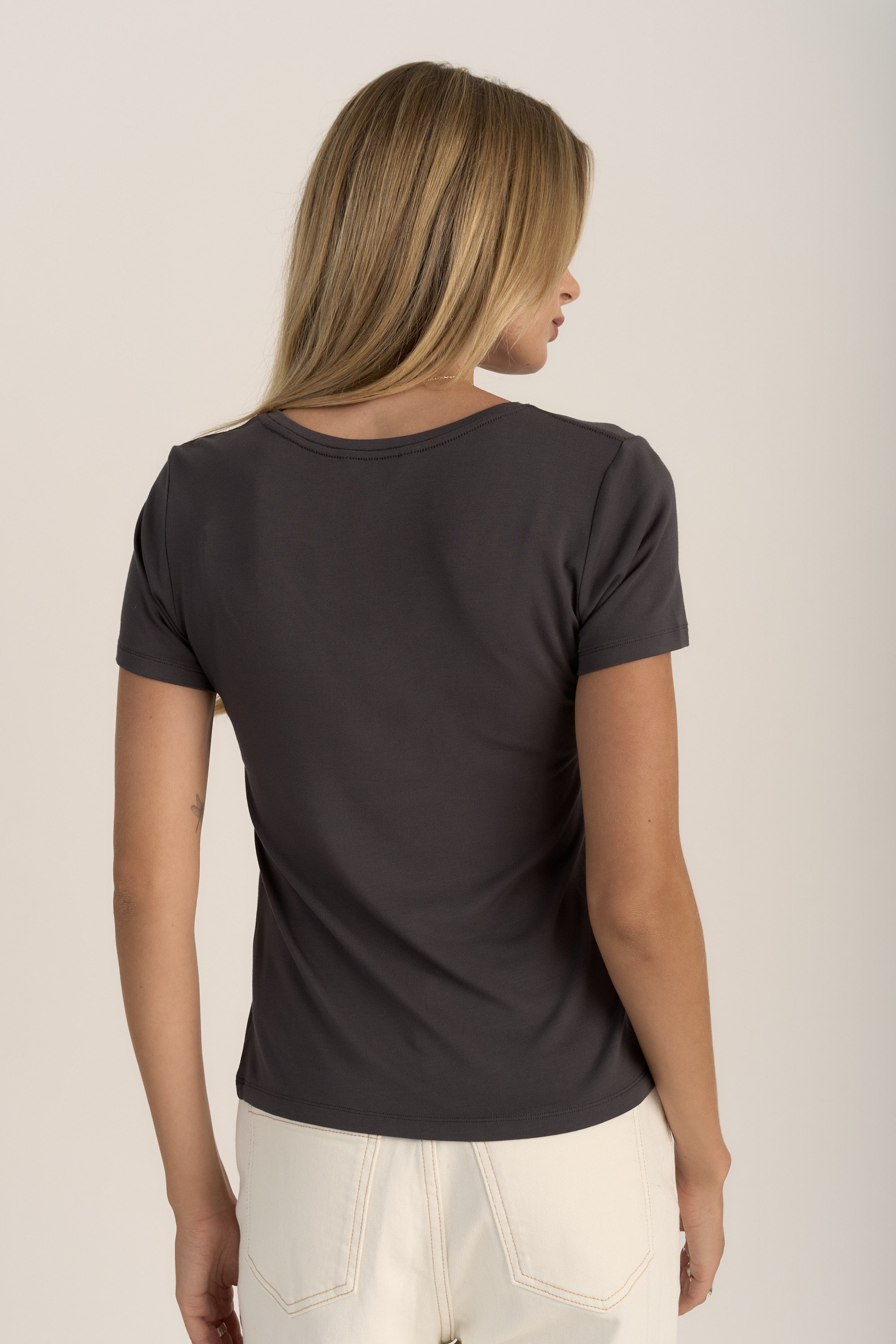 A woman with long blonde hair stands with her back to the camera, wearing the Max Riley Short Sleeve V Neck Tee and light-colored pants against a plain, light background.