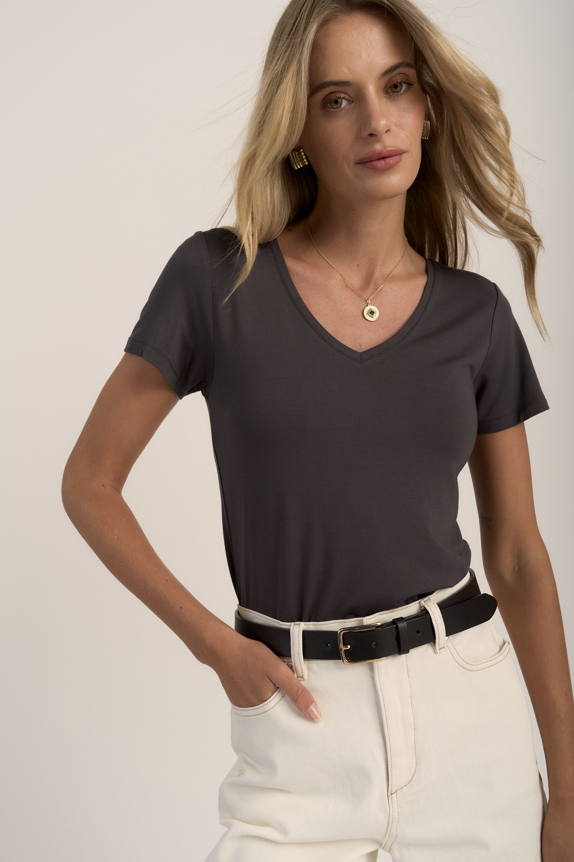 A woman with long blonde hair wears the Max Riley Short Sleeve V Neck Tee, light high-waisted jeans with a black belt, and gold jewelry. She stands with one hand in her pocket against a neutral background.