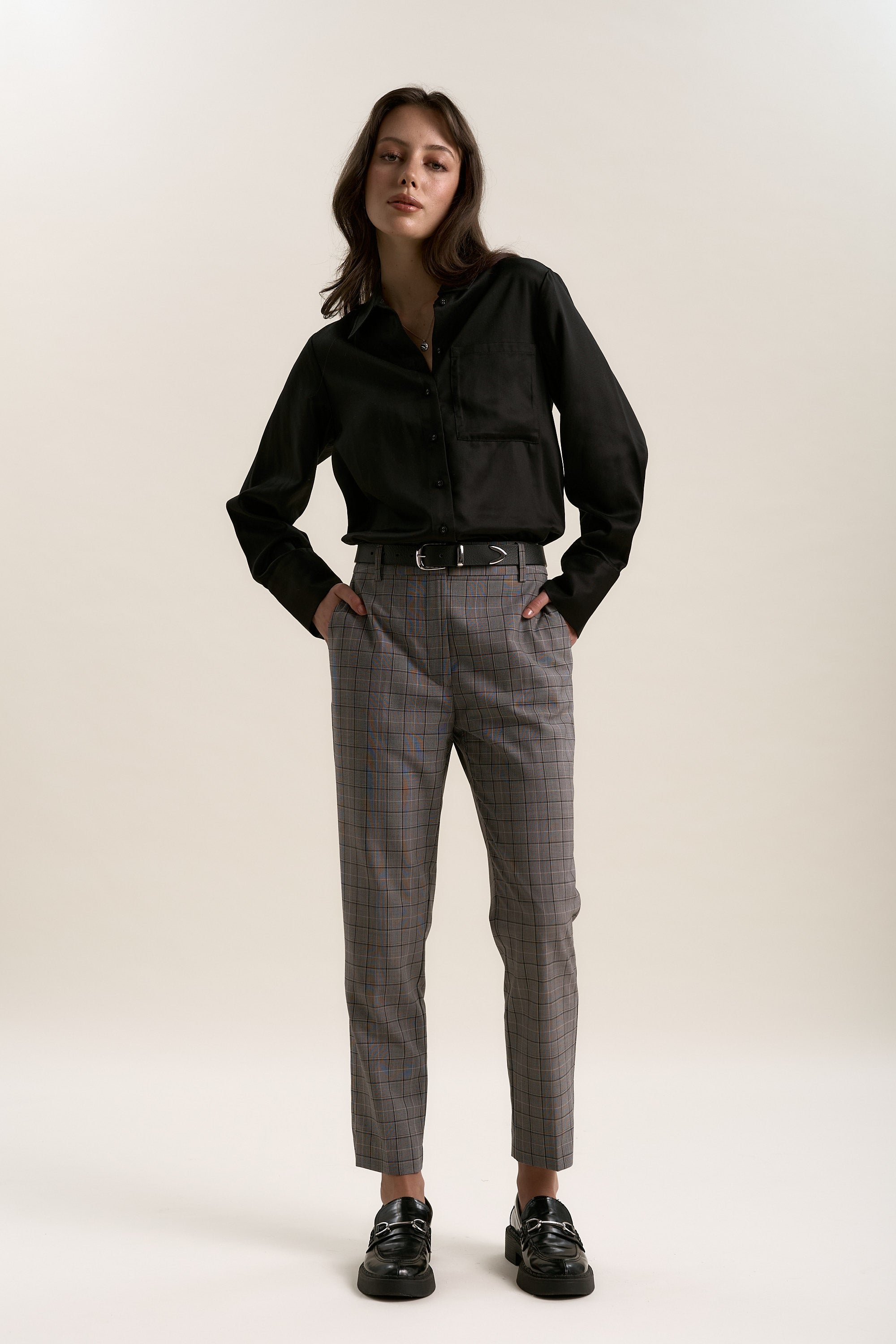 A woman stands against a plain background, hands in pockets, wearing Max's The Wool Blend Pant—a plaid, tapered office style with belt—paired with a black button-up shirt and black loafers. She looks confidently at the camera.