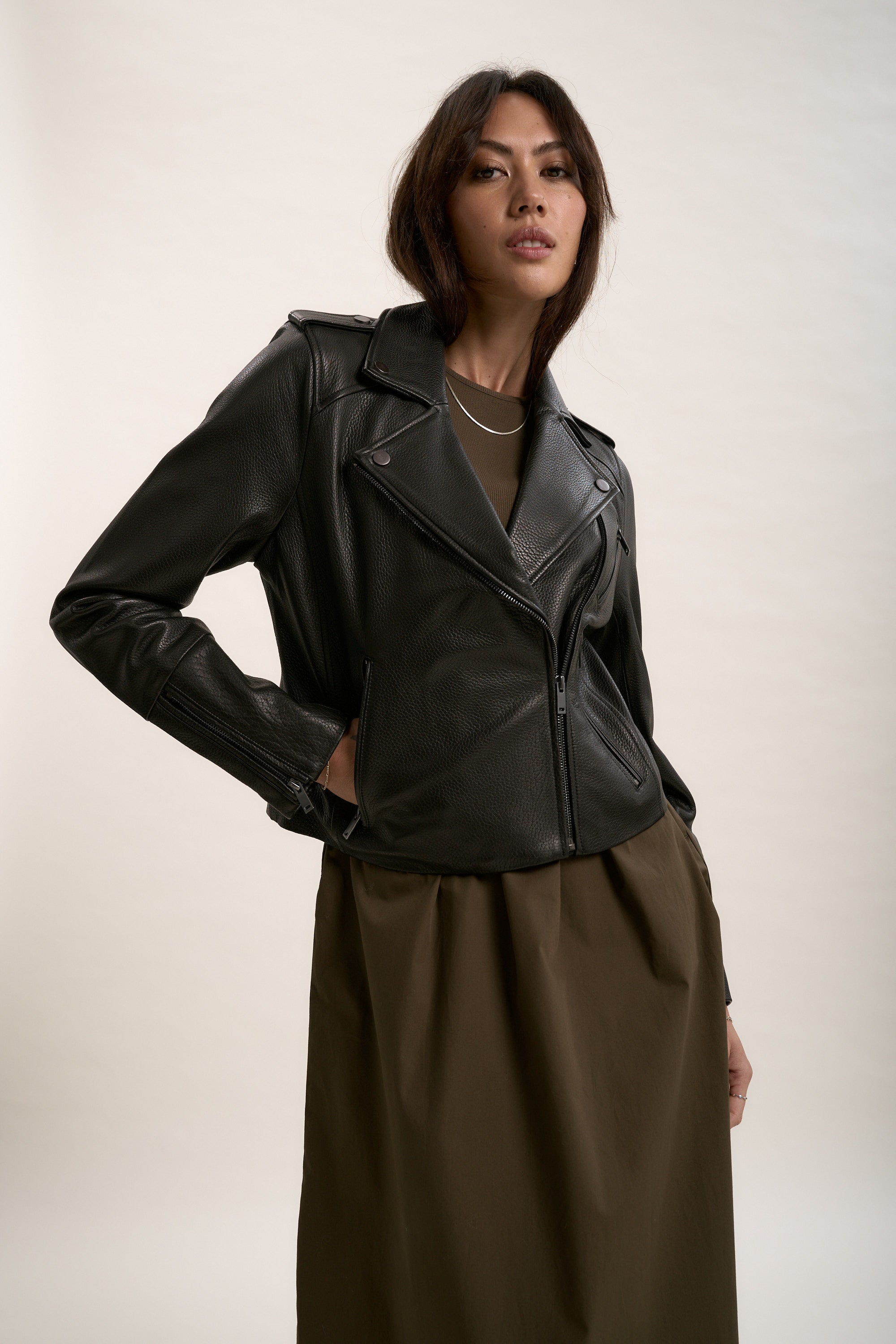 A woman with medium-length brown hair looks confidently at the camera, wearing Max’s The Perfect Leather Biker Jacket in premium black textured leather over an olive green dress, set against a plain light background.