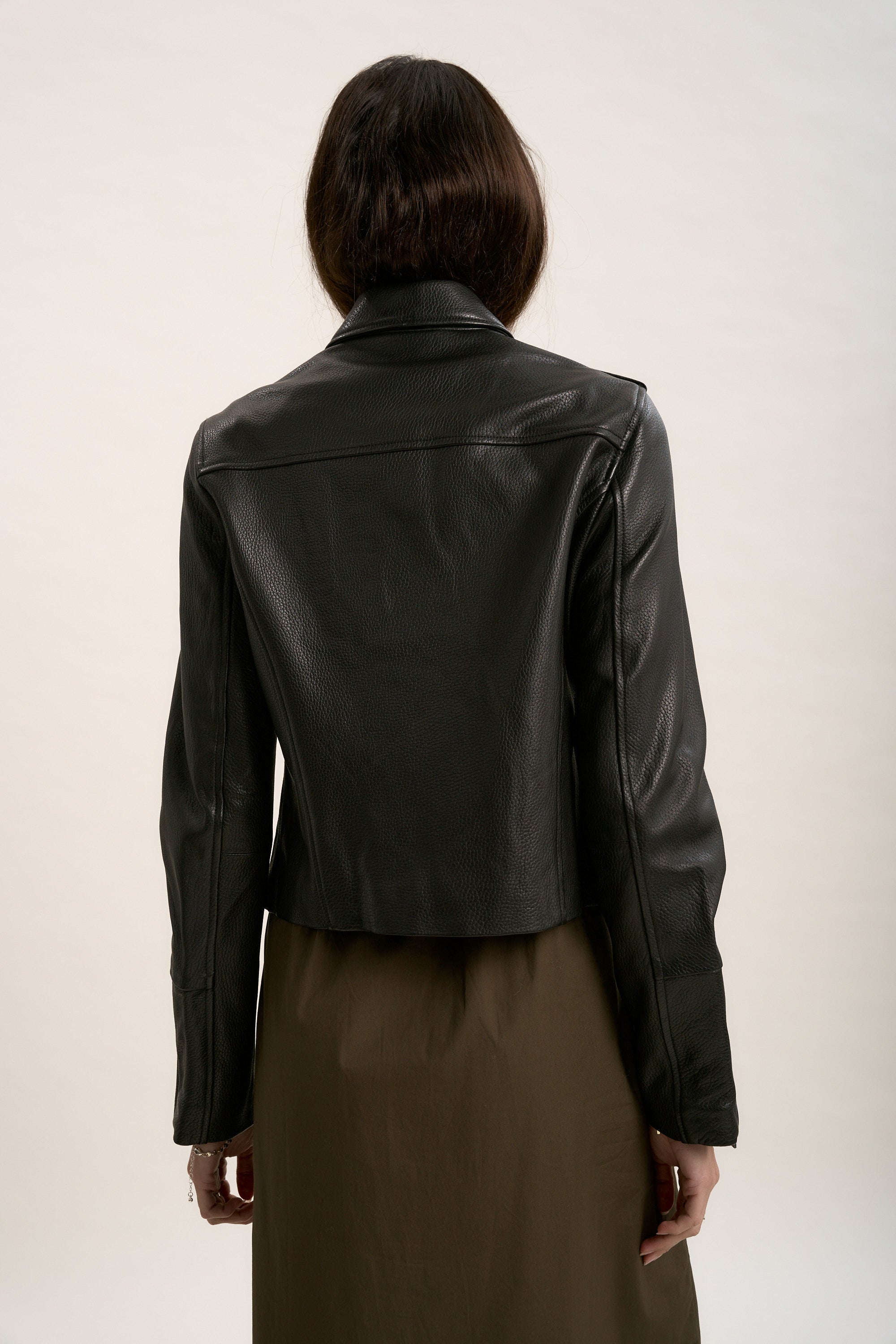 A person with long dark hair, seen from behind, wears Max's The Perfect Leather Biker Jacket—crafted from premium textured leather—over an olive green dress, standing out among women’s wardrobe staples against a plain light background.