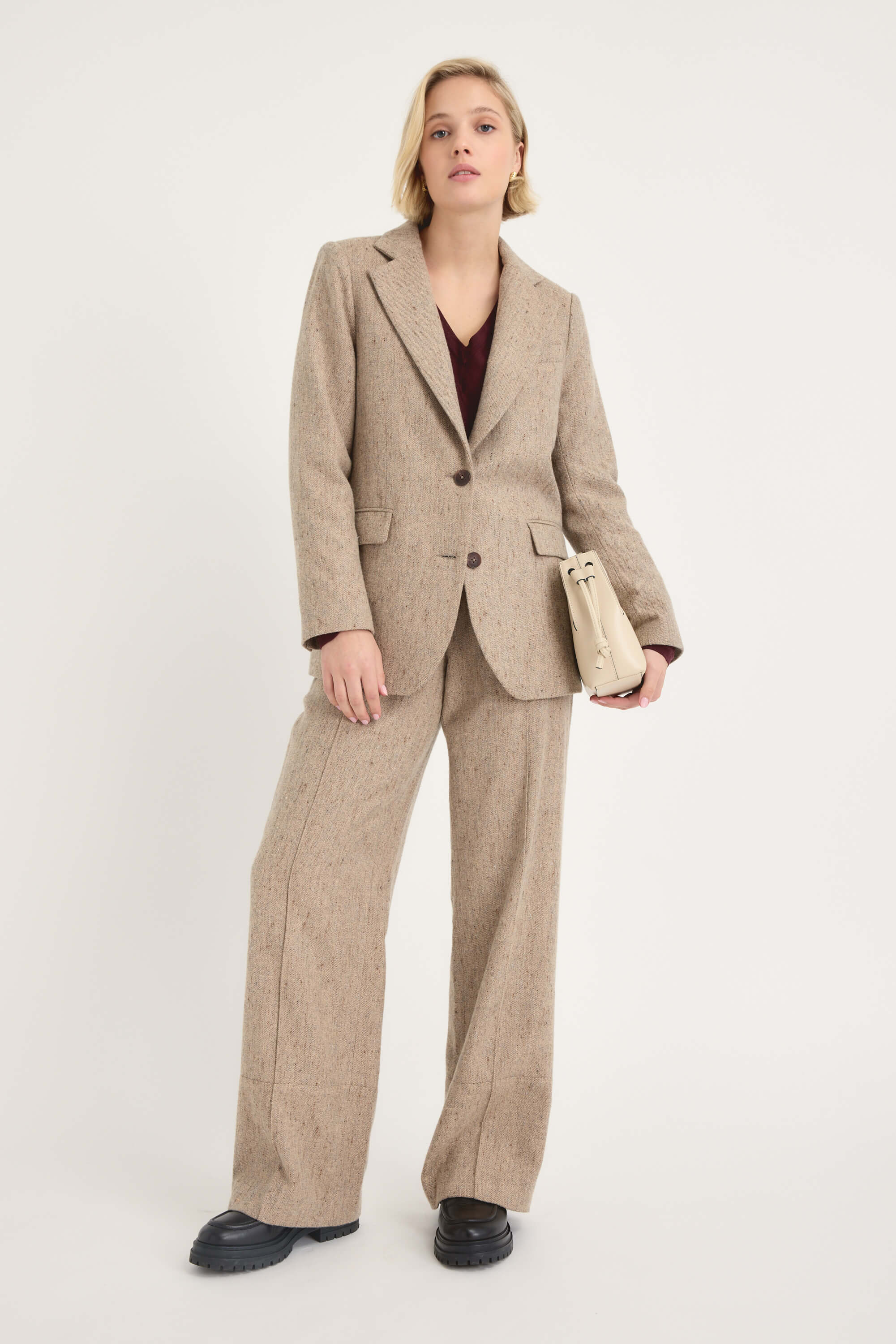A person with short blonde hair wears the Max Luna Wide Leg Pant in tan with a dark top, holds a light-colored handbag, and stands against a plain white background.