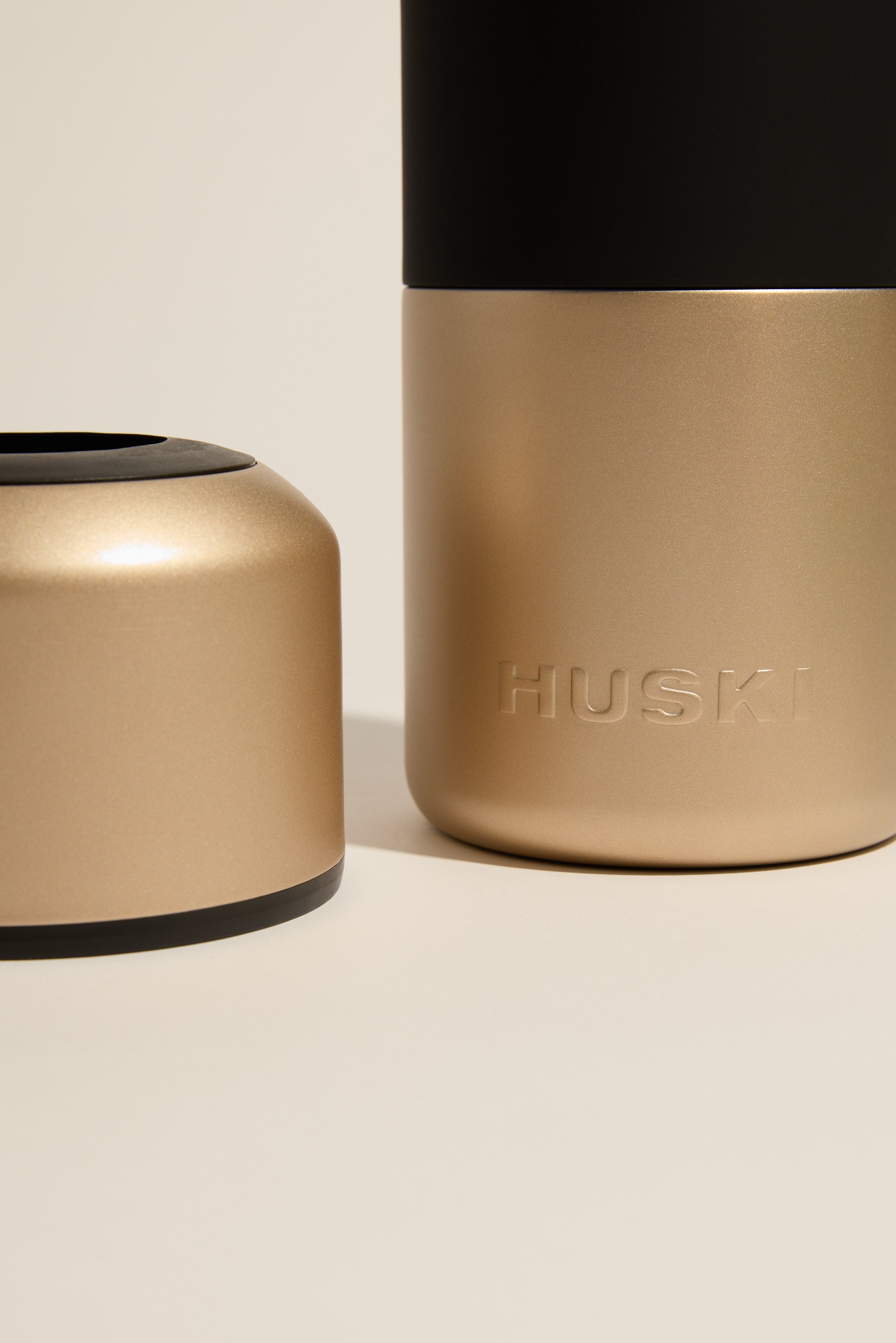 A close-up of the Huski Wine Cooler, a premium stainless steel wine cooler by Huski in metallic gold and black, with "Huski" embossed on the side. The insulated bottle’s cap rests beside it on a light beige surface.