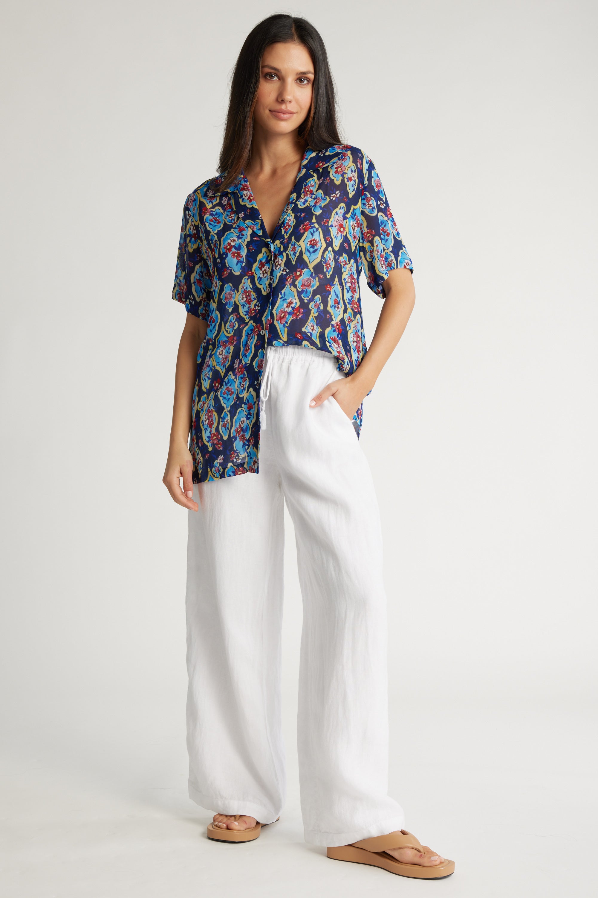 A woman with long dark hair poses against a plain background in the Max Brianna Short Sleeve Shirt, styled with white wide-leg pants and beige sandals. Her left hand is in her pocket, showcasing perfect summer fashion.