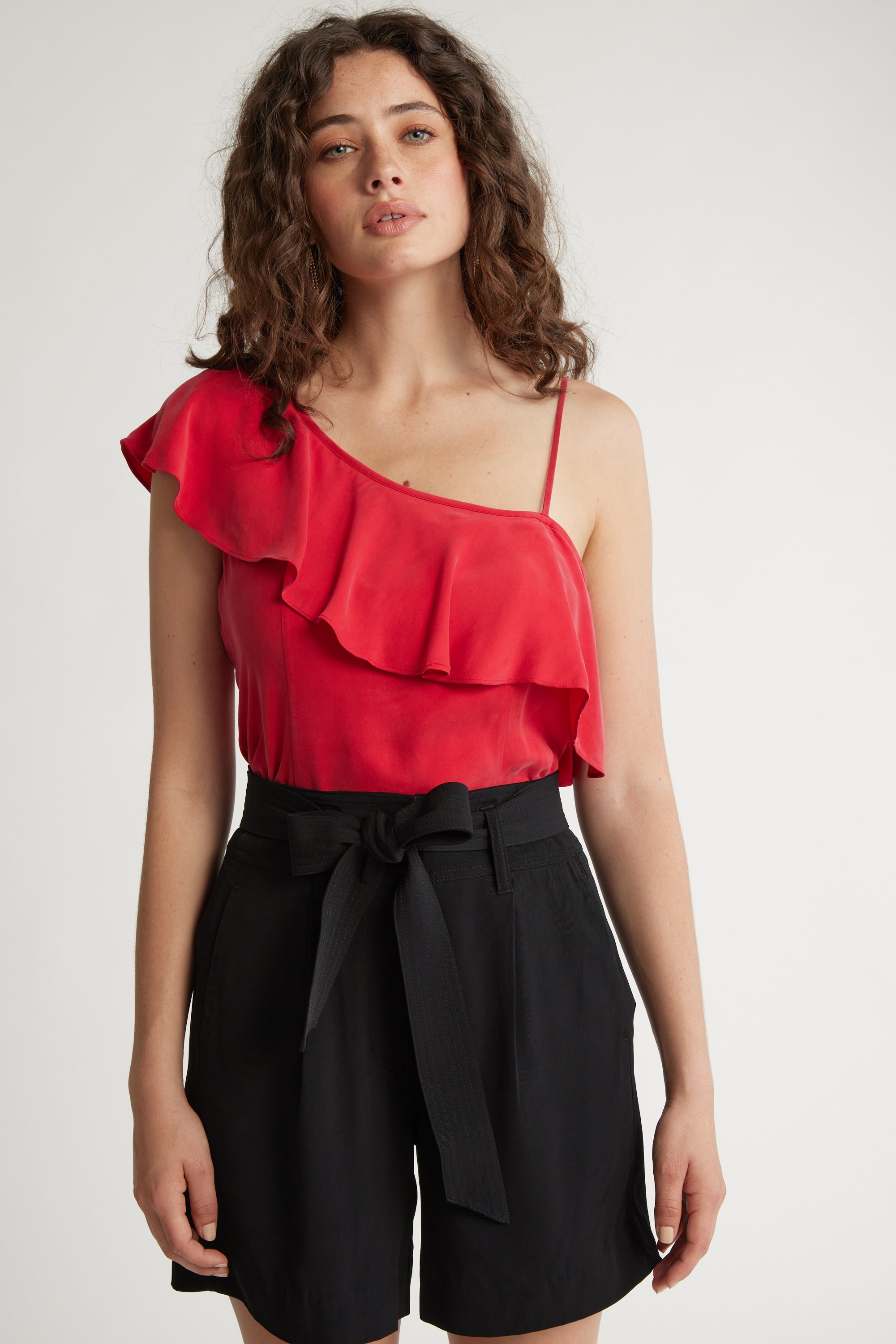 A woman with curly brown hair wears the Max Molly Asymmetric Ruffle Top in red, paired with high-waisted black shorts with a tied belt, standing against a plain light background.