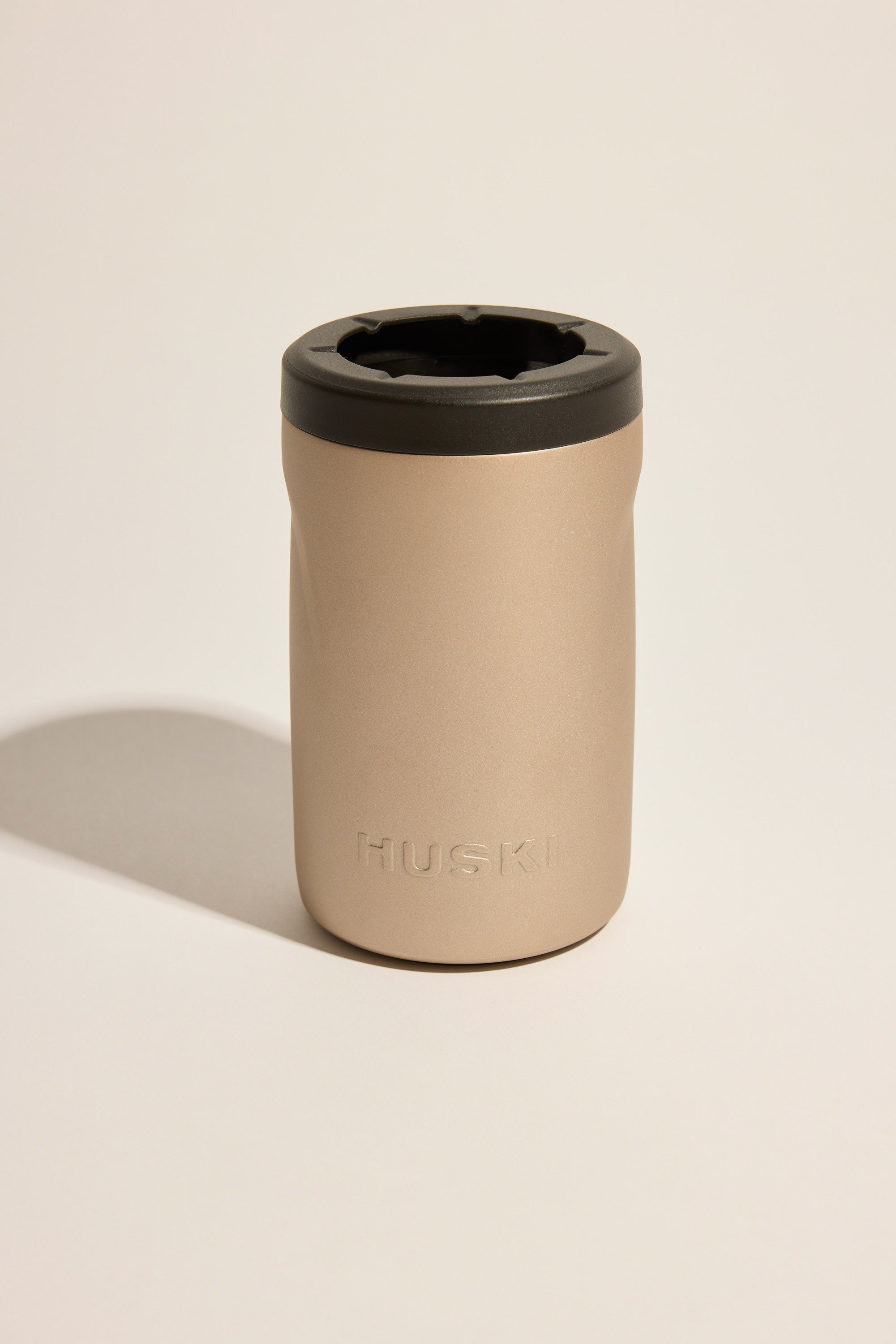 The Huski Beer Cooler by Huski is a beige insulated tumbler with a black lid, featuring the HUSKI logo. It stands upright on a light neutral surface with a soft shadow.