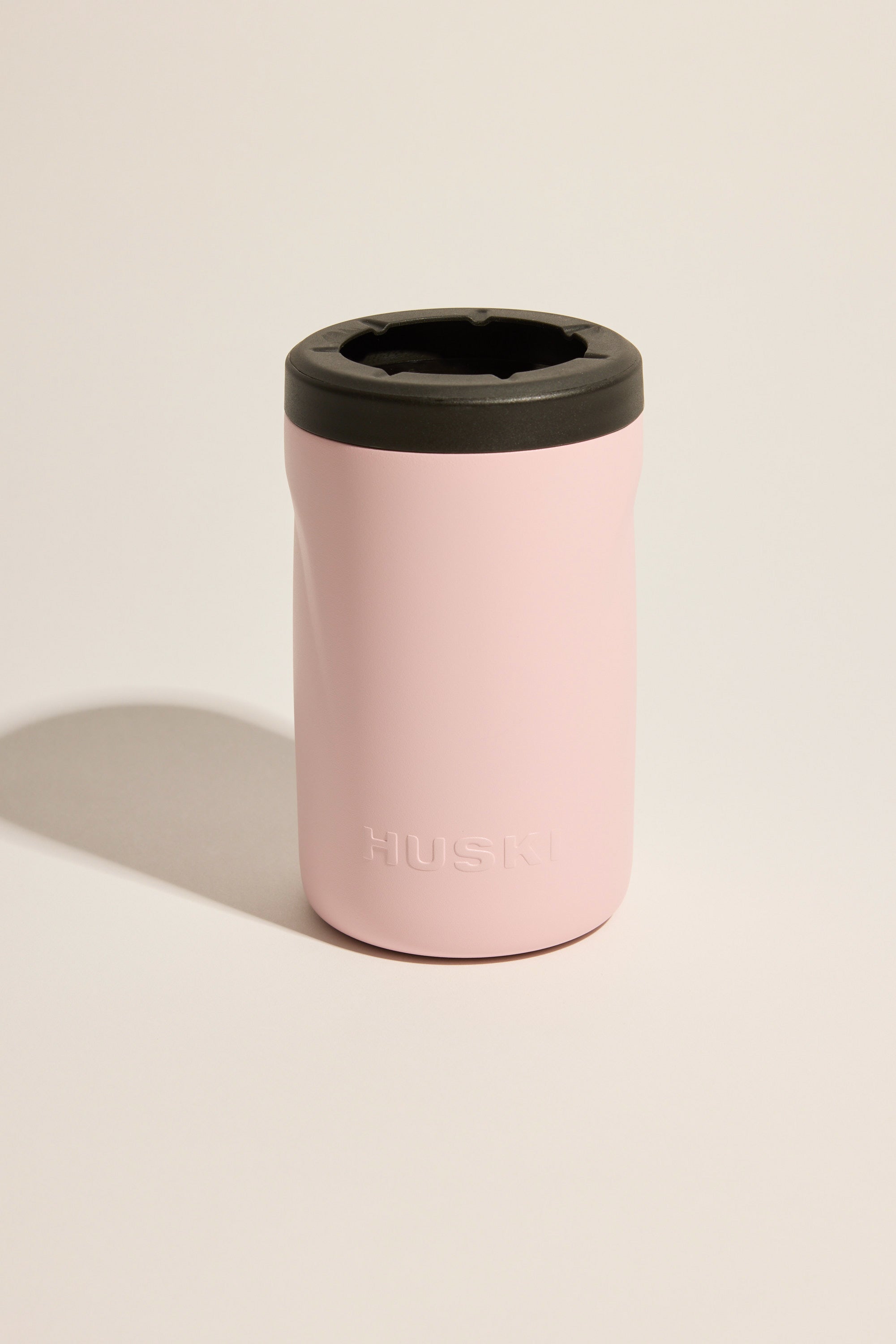 A light pink Huski Beer Cooler with a black lid rests on a neutral background. "HUSKI" is embossed on the front of the stainless steel cooler from Huski.