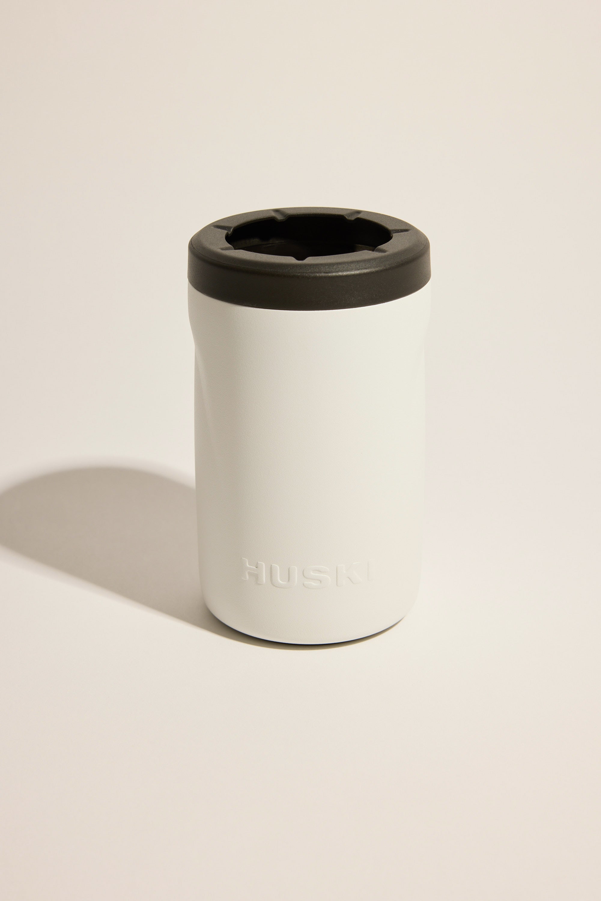 A white Huski Beer Cooler by Huski, made from triple-insulated stainless steel with a black lid and branded embossing, stands upright on a light surface with a gentle shadow.