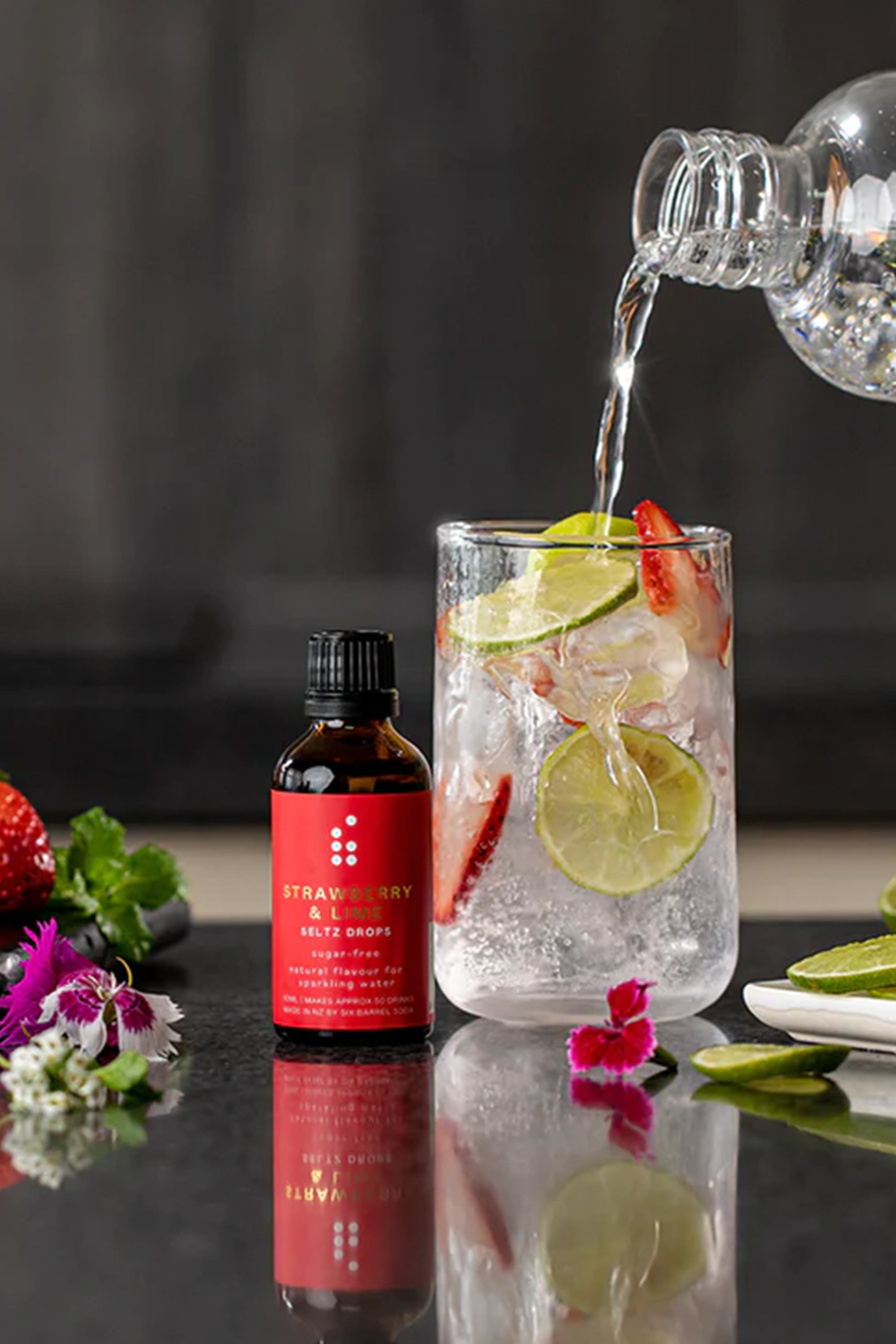 A bottle of Six Barrel Soda Seltz Drops by Six Barrel Soda Company sits beside a glass with ice, lime, and strawberry as sparkling water is poured in. Fresh fruit and flowers are arranged nearby on a dark surface.