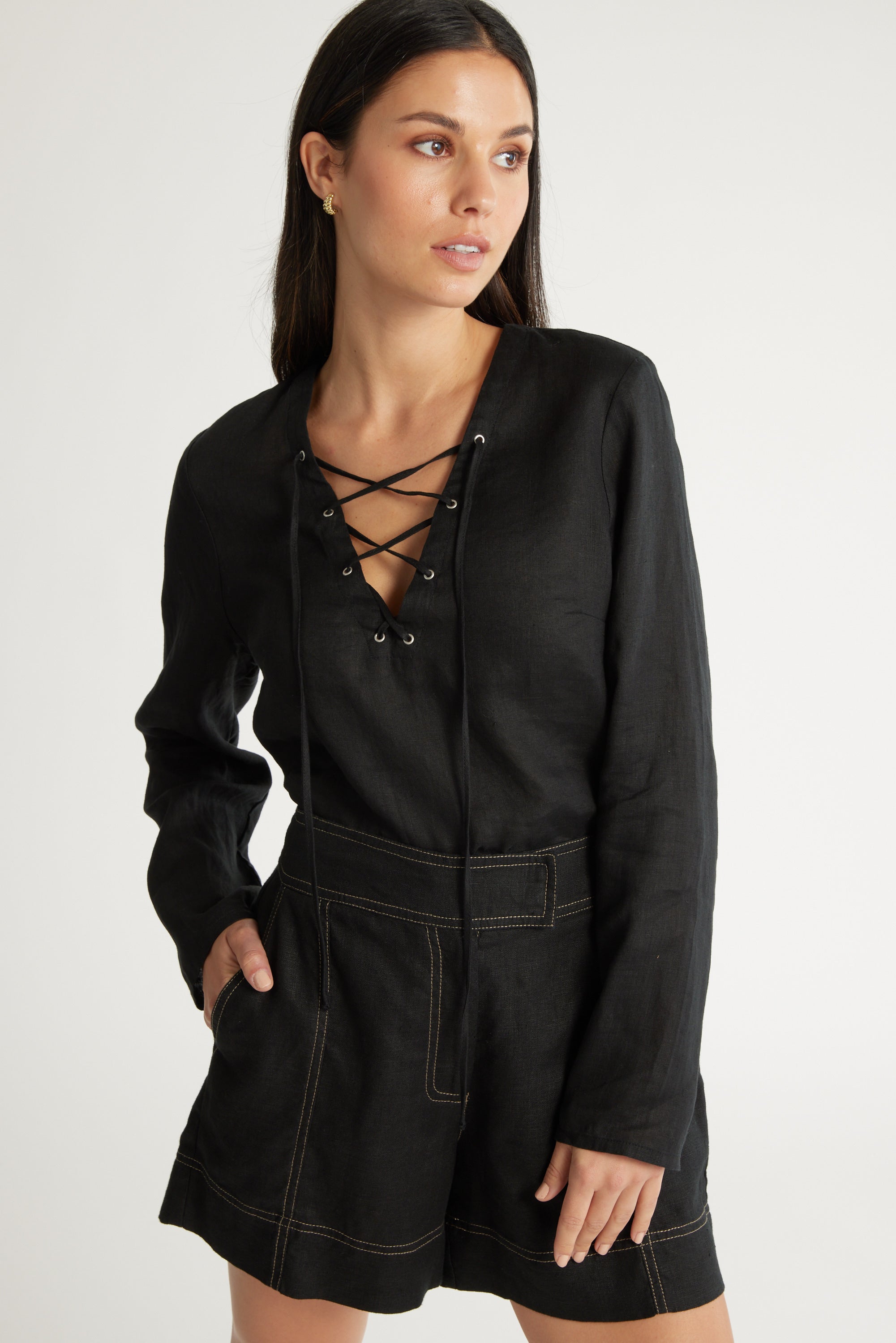 A woman stands with one hand in her pocket, wearing the Max Lucia Tie Detail Top—a black long sleeve romper with lace-up tie detail and contrast stitching—looking slightly to the side against a plain background.