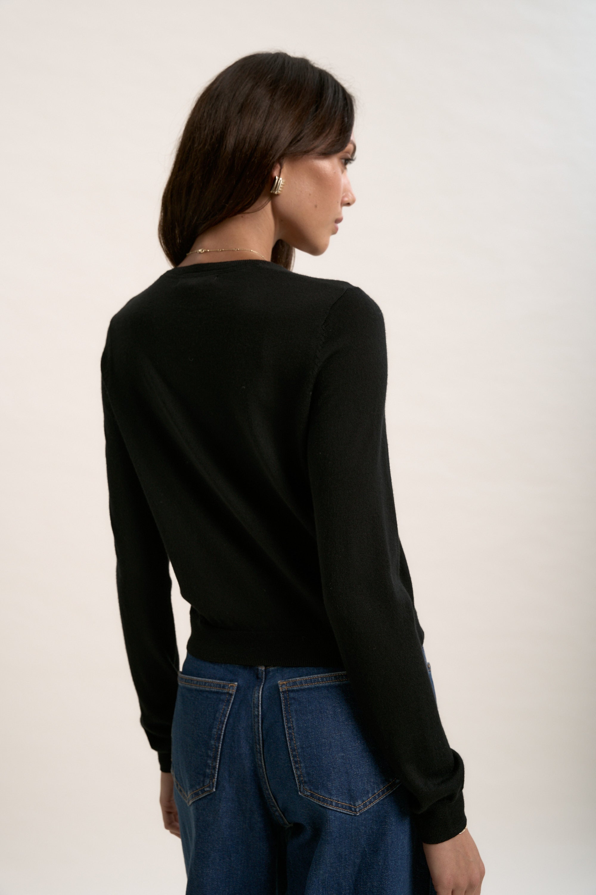 A person with long brown hair, in Max's The Merino Silk Cardigan and blue jeans, stands turned away at an angle against a plain light background.