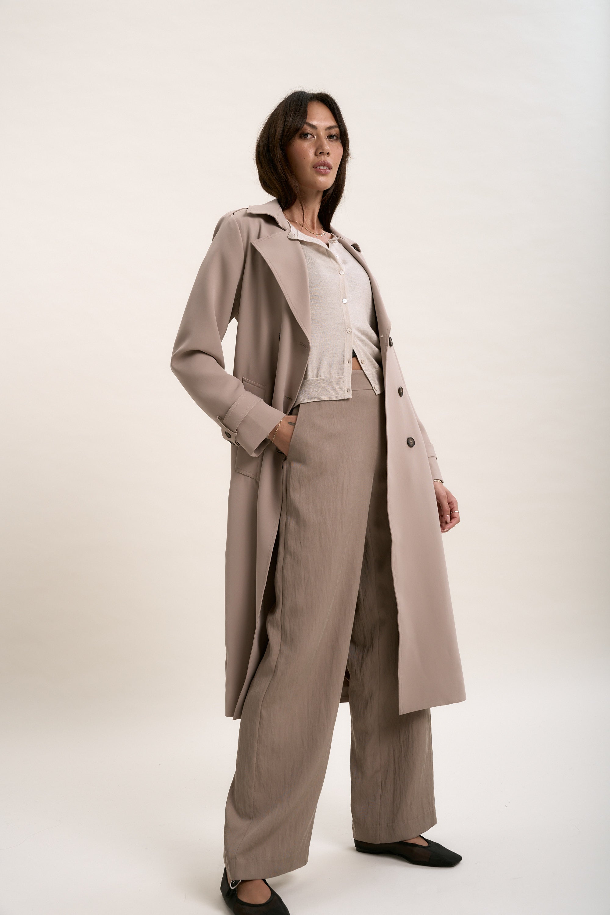 A woman stands confidently in a beige trench coat, light blouse, and wide-leg trousers, layered with Max's The Merino Silk Cardigan. Hand in pocket, she gazes upward against a plain light background.